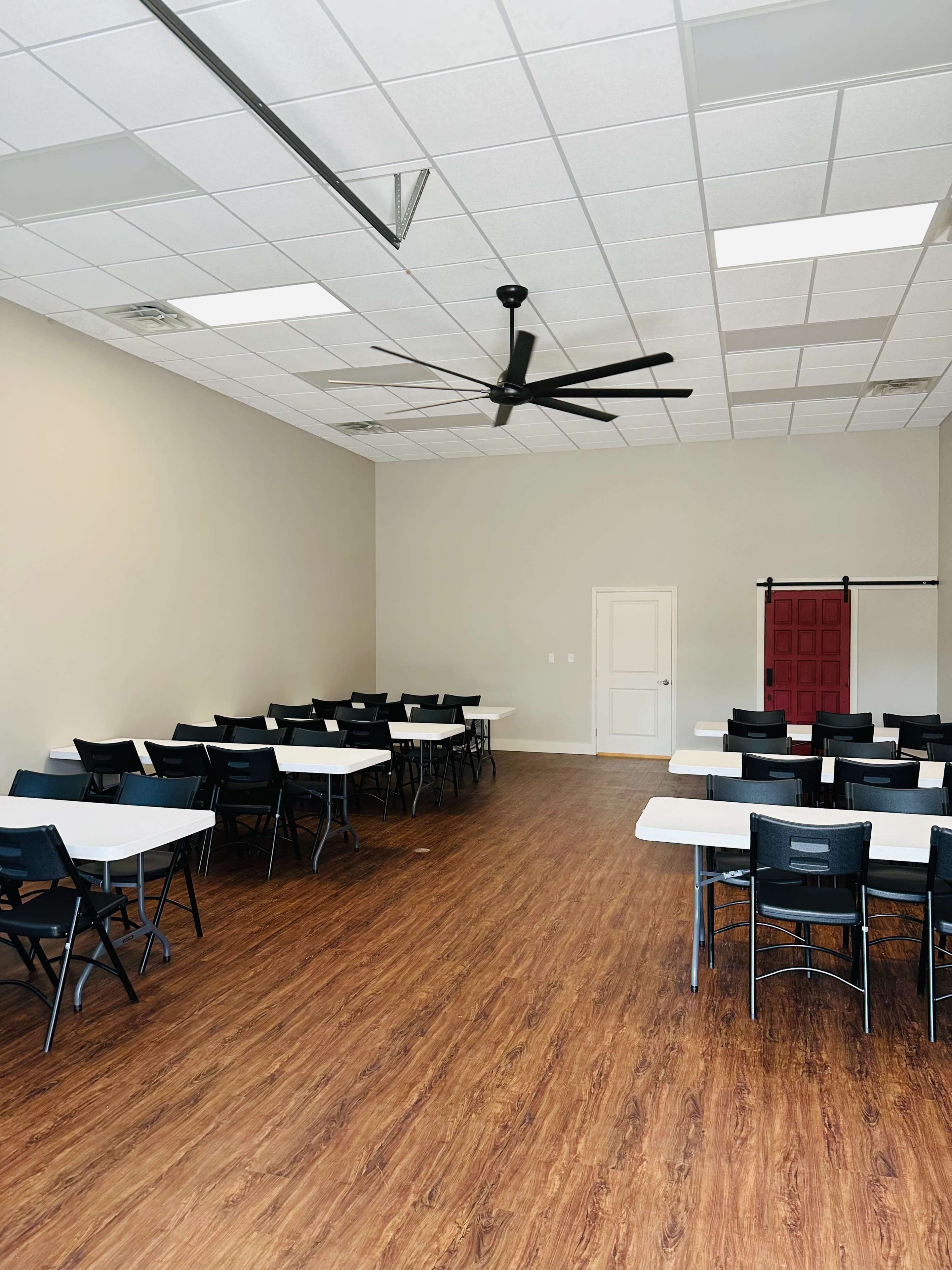 A spacious meeting room with rows of rectangular tables and black chairs, featuring a ceiling fan and a red door at the back.