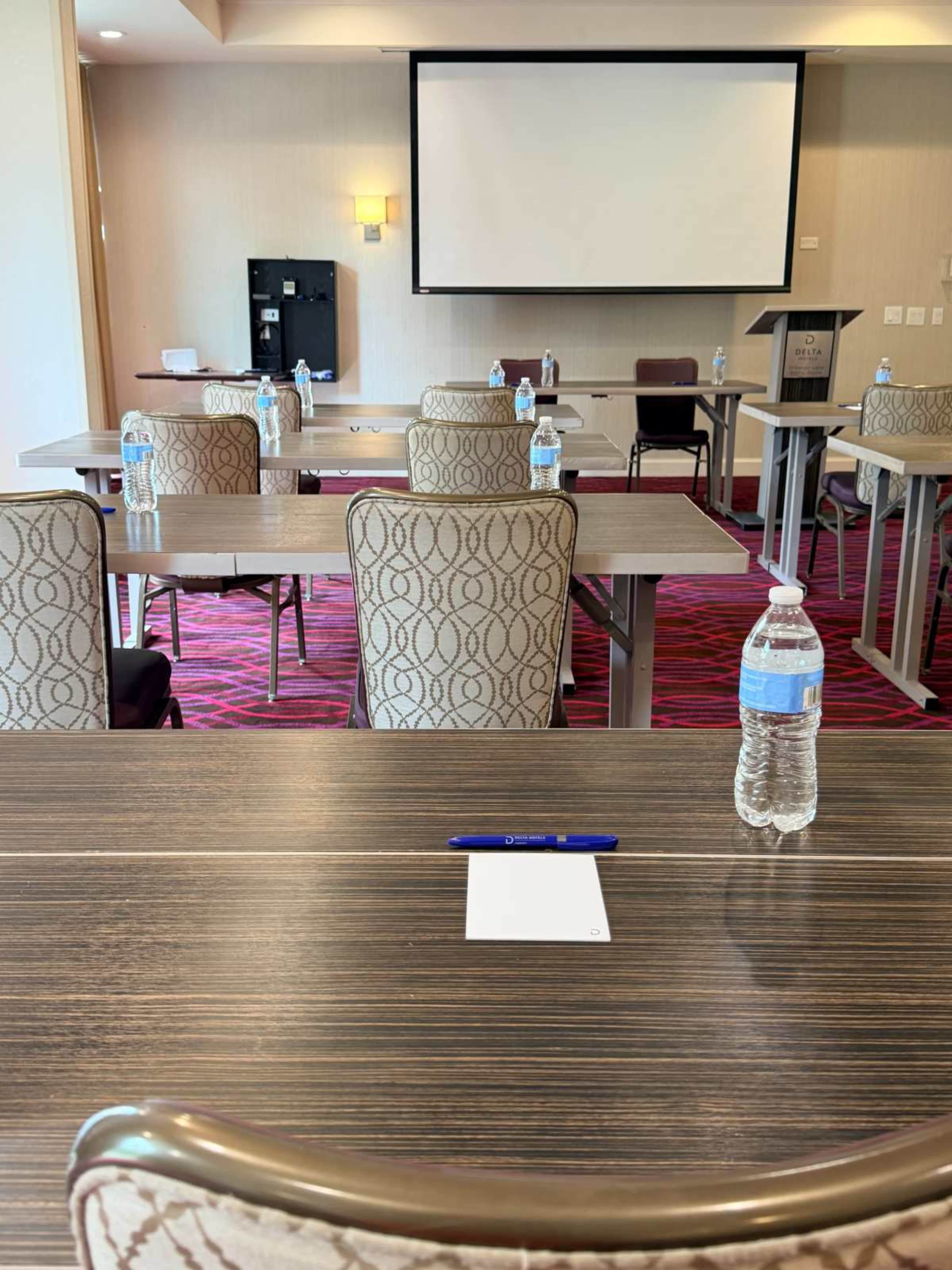 The image shows a conference room set up with tables and chairs, each equipped with a bottle of water and a notepad, facing a presentation screen.