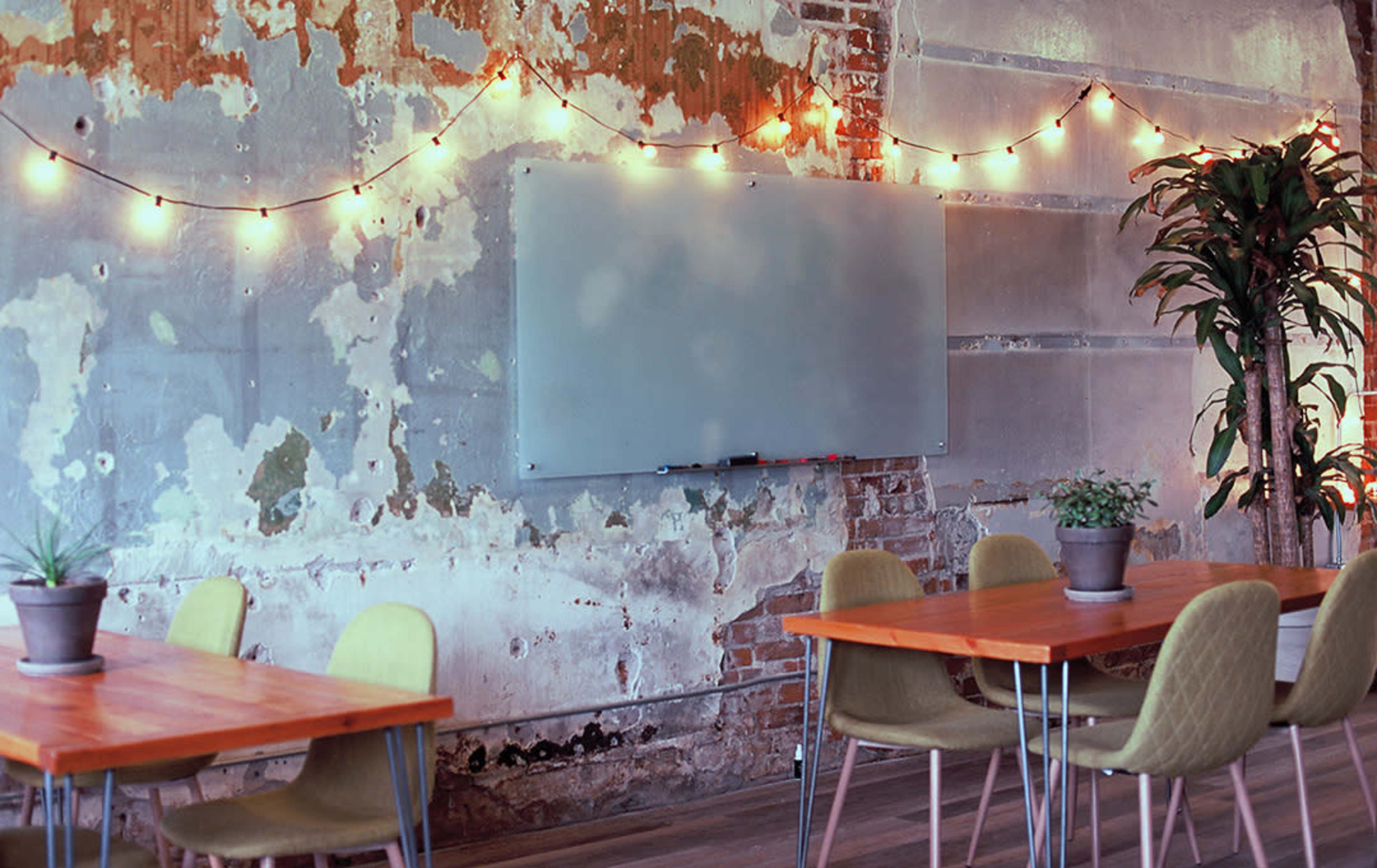 A rustic interior with a weathered wall, string lights, a blank whiteboard, and several tables surrounded by chairs, along with potted plants.