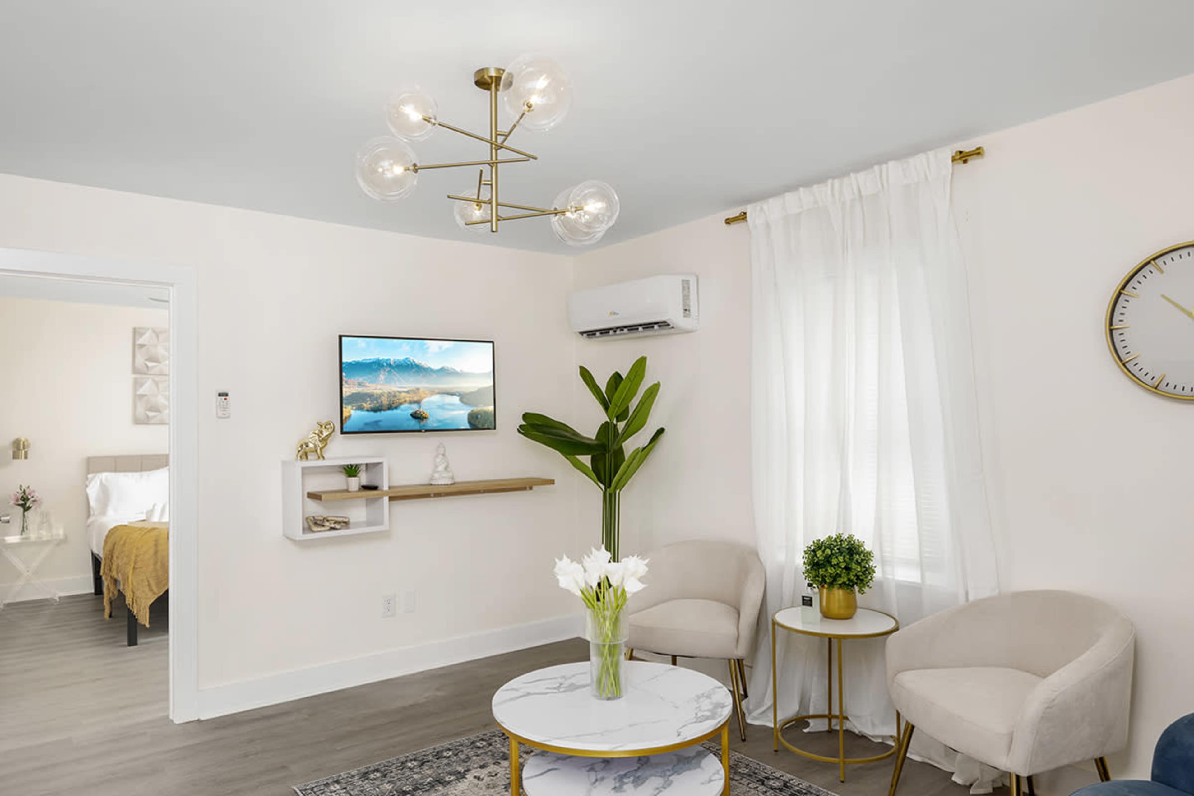 A modern living room features two light-colored chairs, a round marble coffee table, a television mounted on the wall, and a large potted plant near a window with sheer curtains.