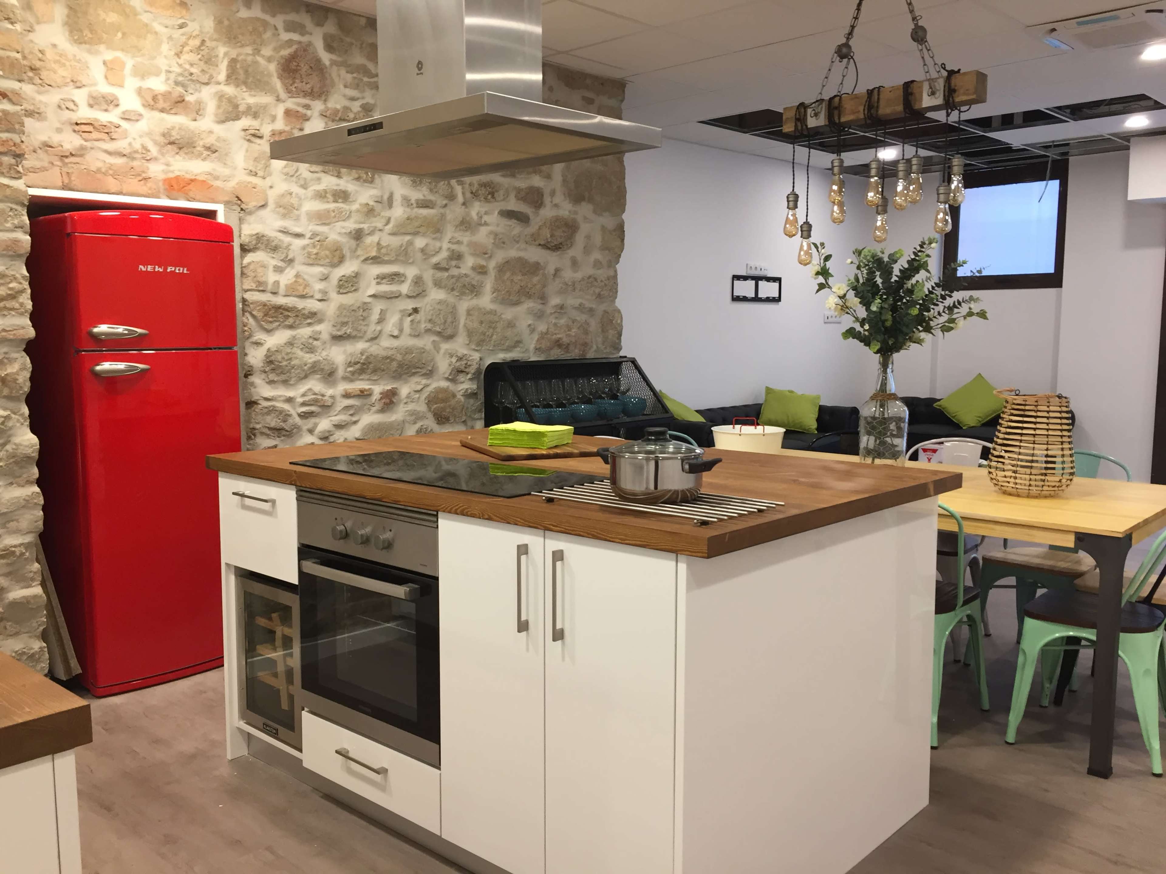 A modern kitchen with a red refrigerator, an island with a cooktop, and a dining area featuring a wooden table and green chairs.