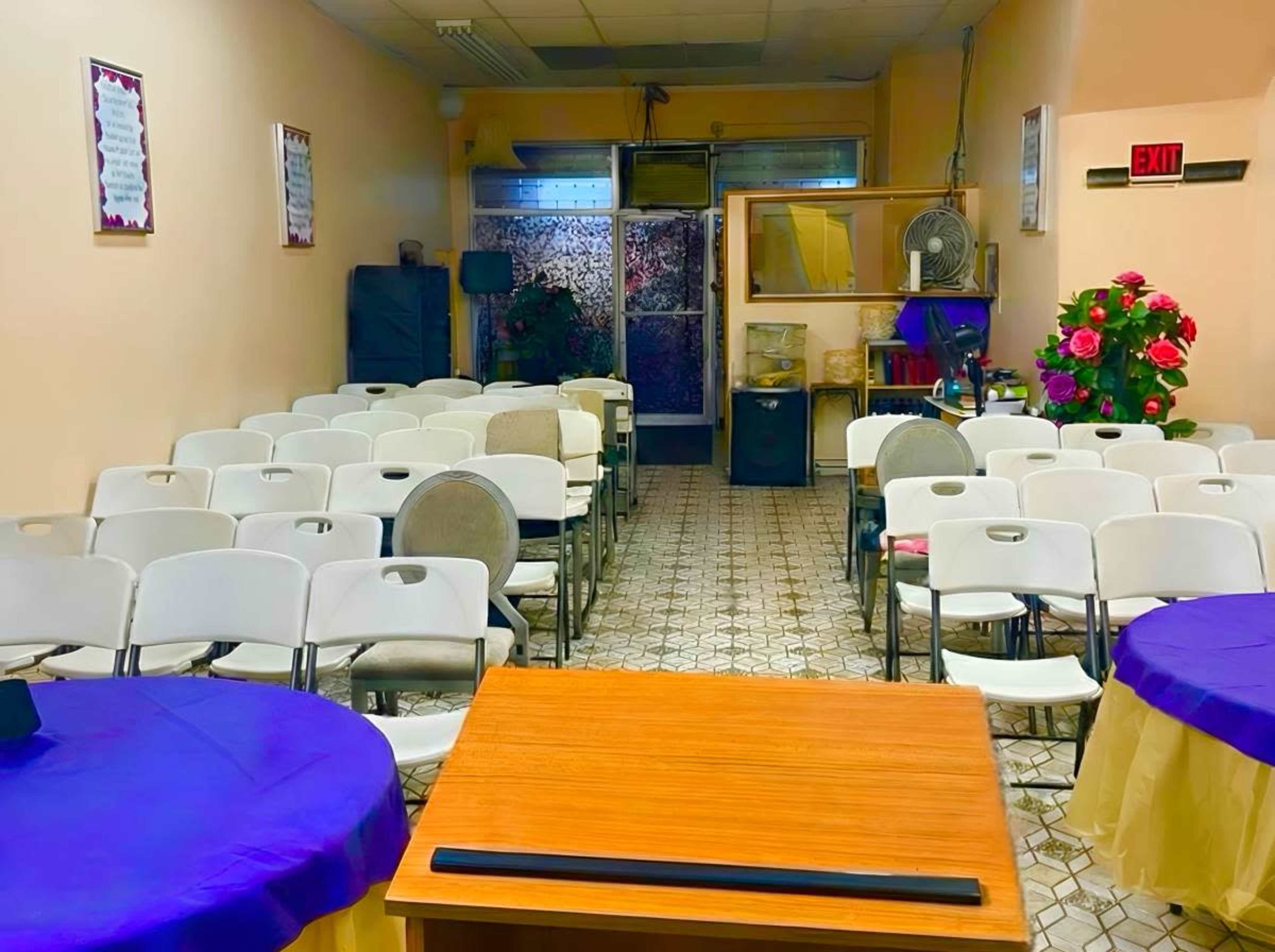 The image shows a partially filled room with rows of white chairs arranged around a wooden table, featuring decorations and a floral display in the corner.