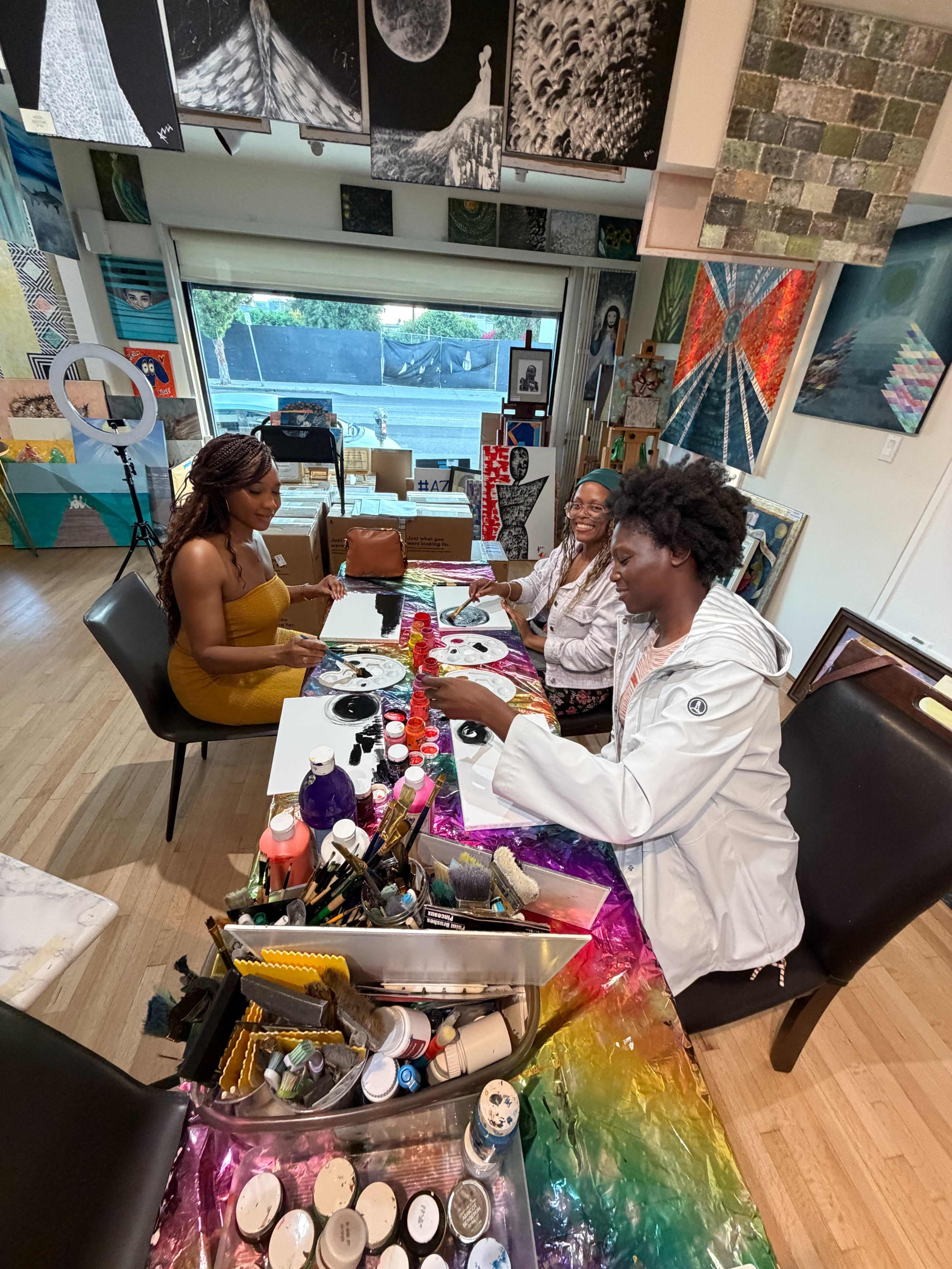 Three women are engaged in a painting session at a table filled with art supplies, with colorful artworks displayed on the walls behind them.