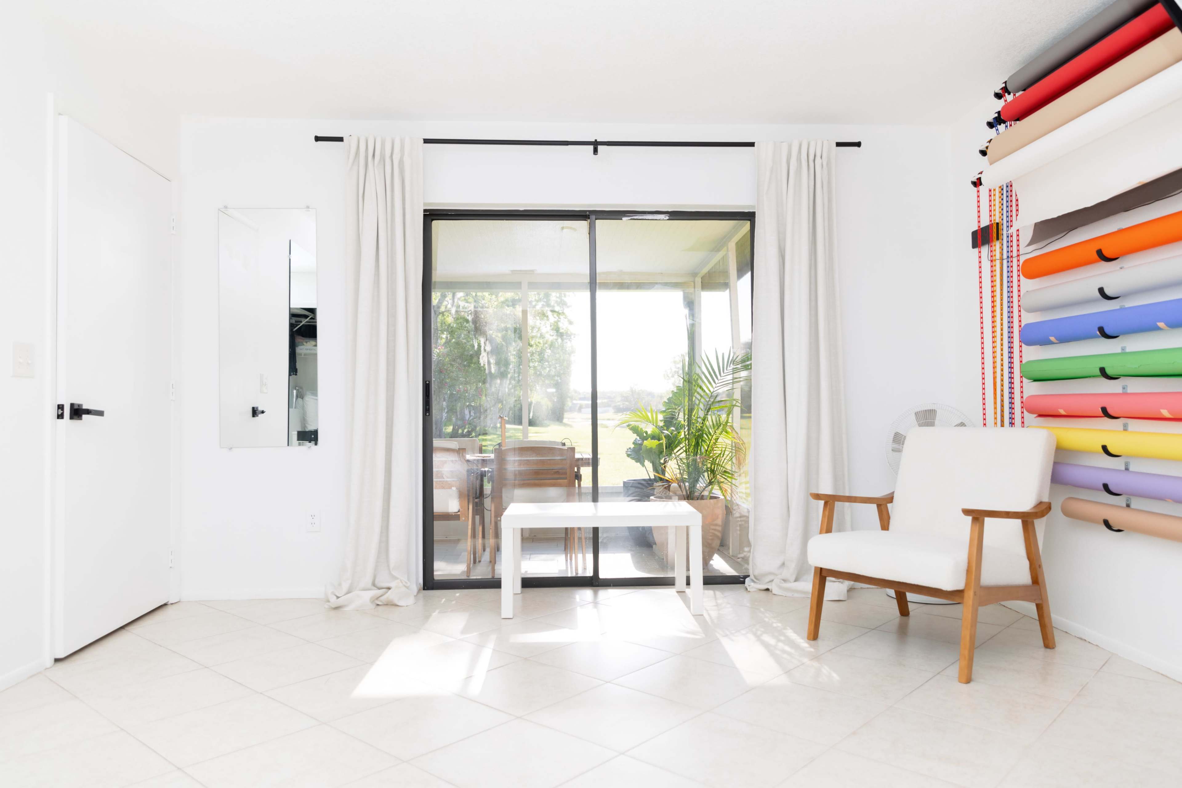 A bright, minimalist room features a chair and a small table near a glass sliding door overlooking a patio and greenery.