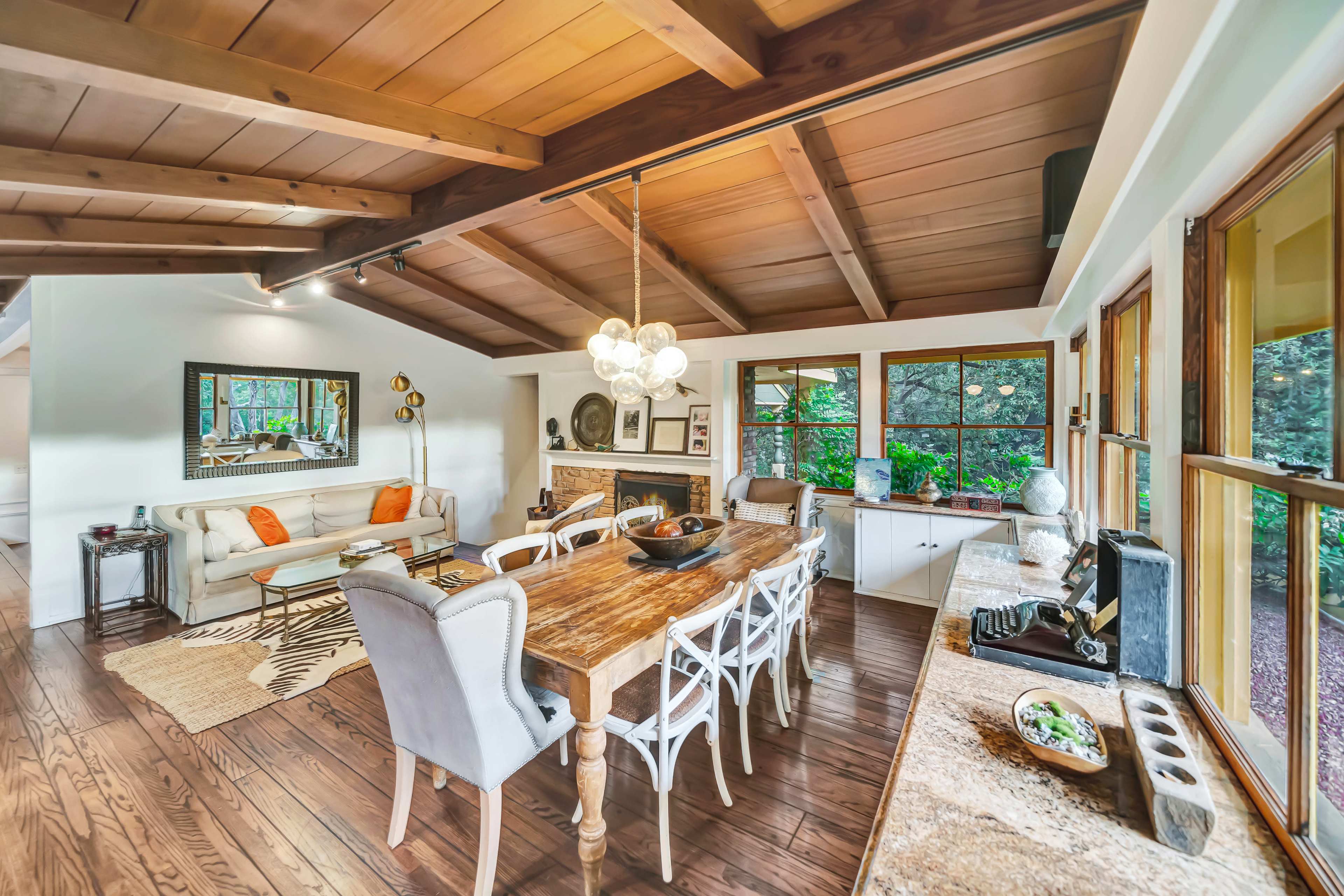 The image shows a dining area featuring a wooden table with light-colored chairs, a cozy living space with a sofa, and large windows overlooking greenery.