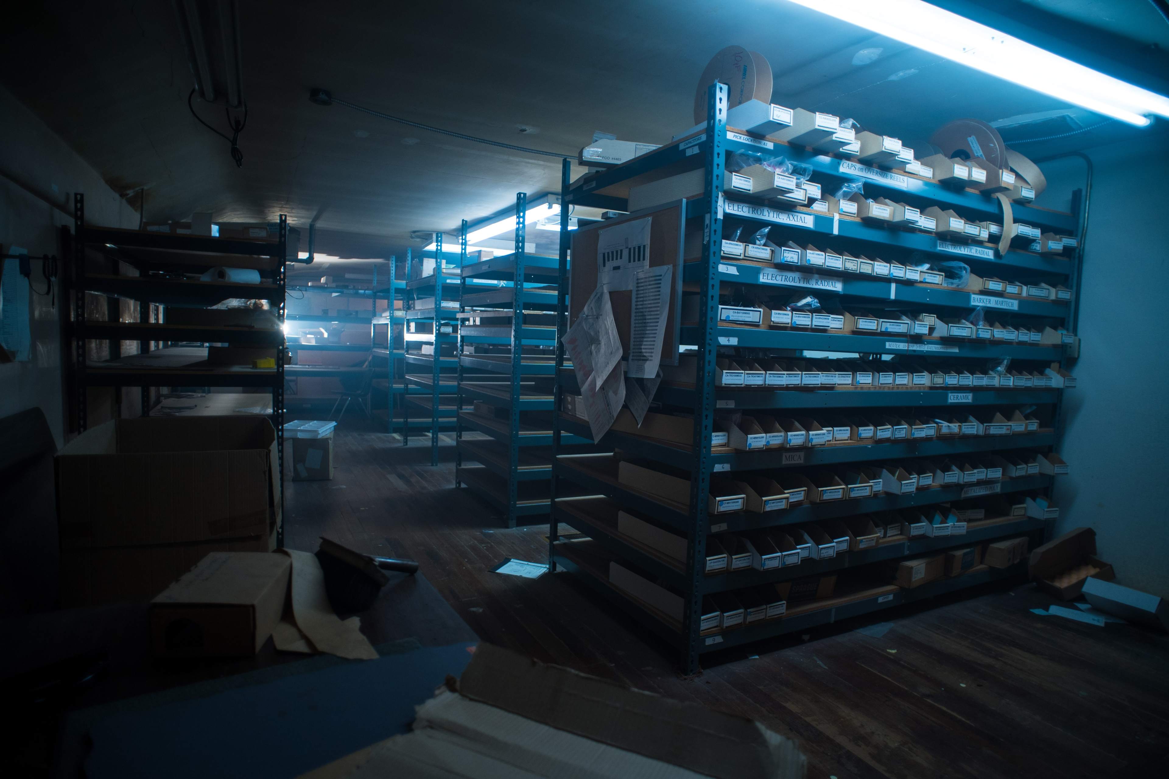 A dimly lit storage room filled with metal shelves stacked with boxes and organized materials, illuminated by overhead fluorescent lights.