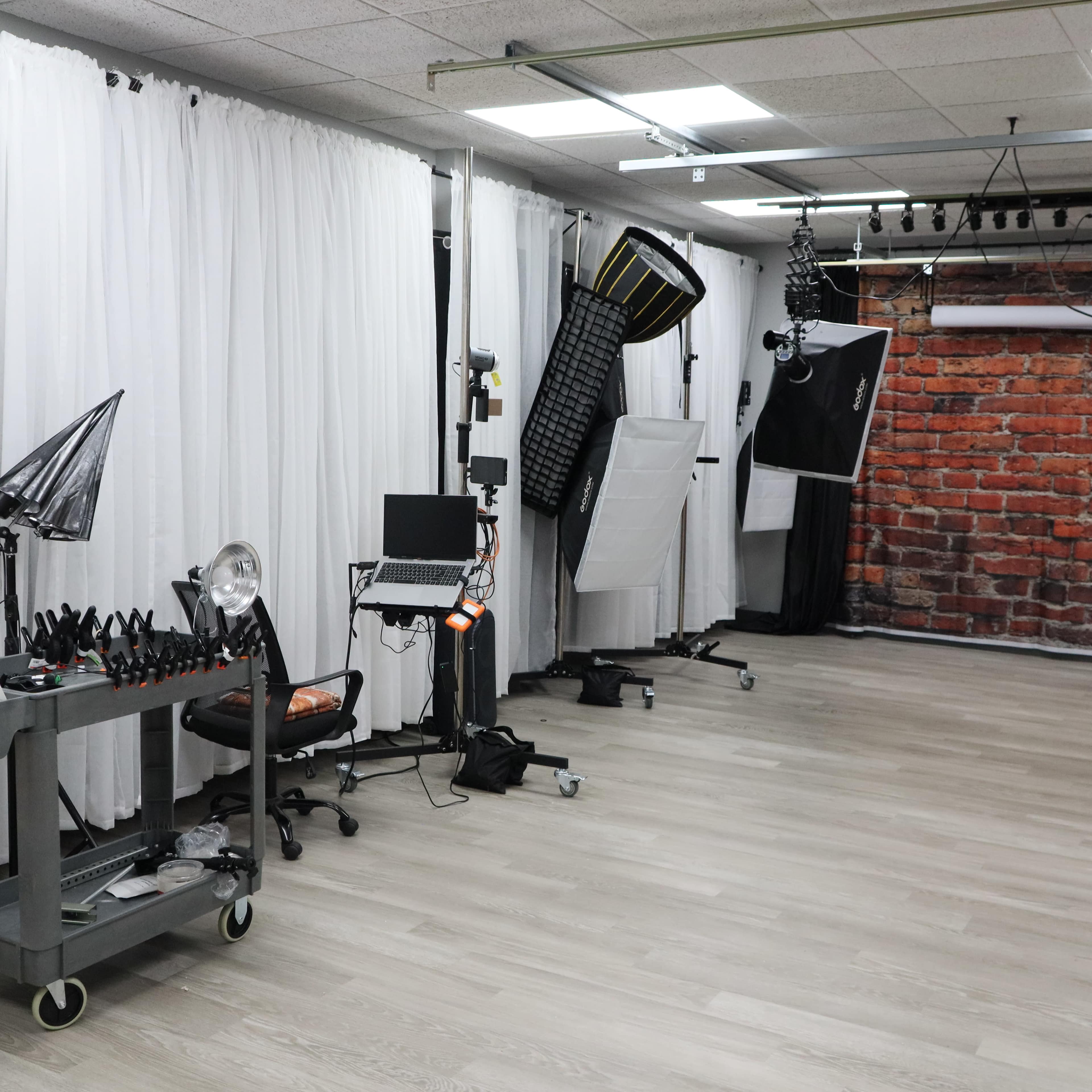 The image shows a photography studio with white curtains, multiple light setups, a computer on a cart, and a brick wall backdrop.