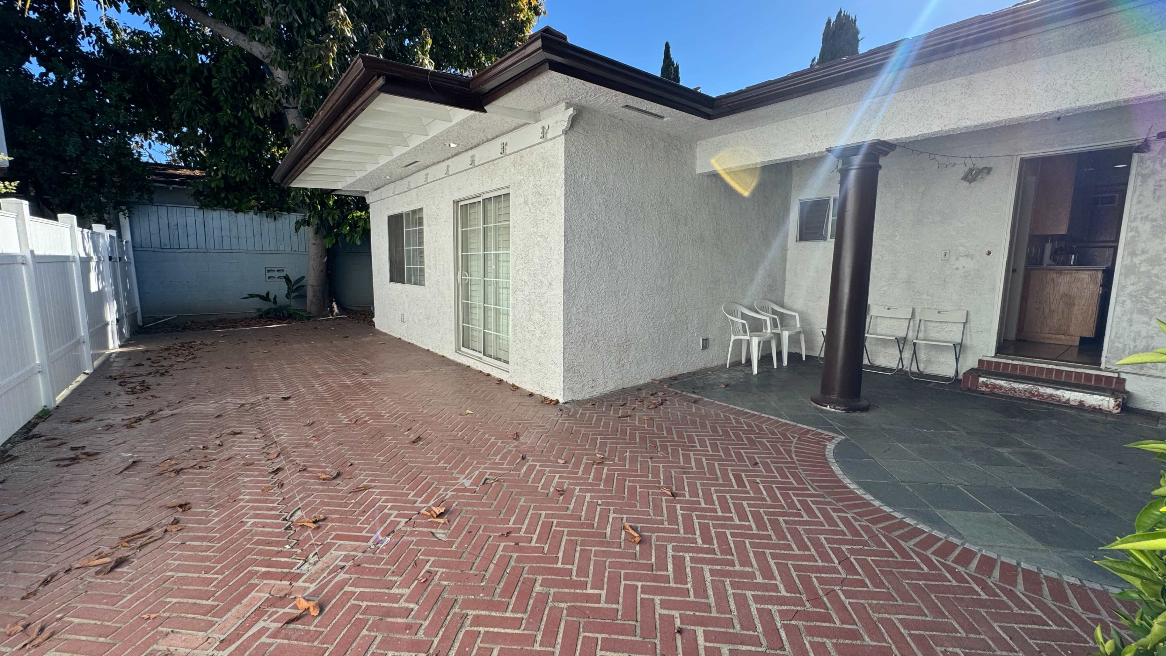 The image shows a patio area with a brick floor and a small covered entrance to a house, featuring a white wall and several plants around the space.