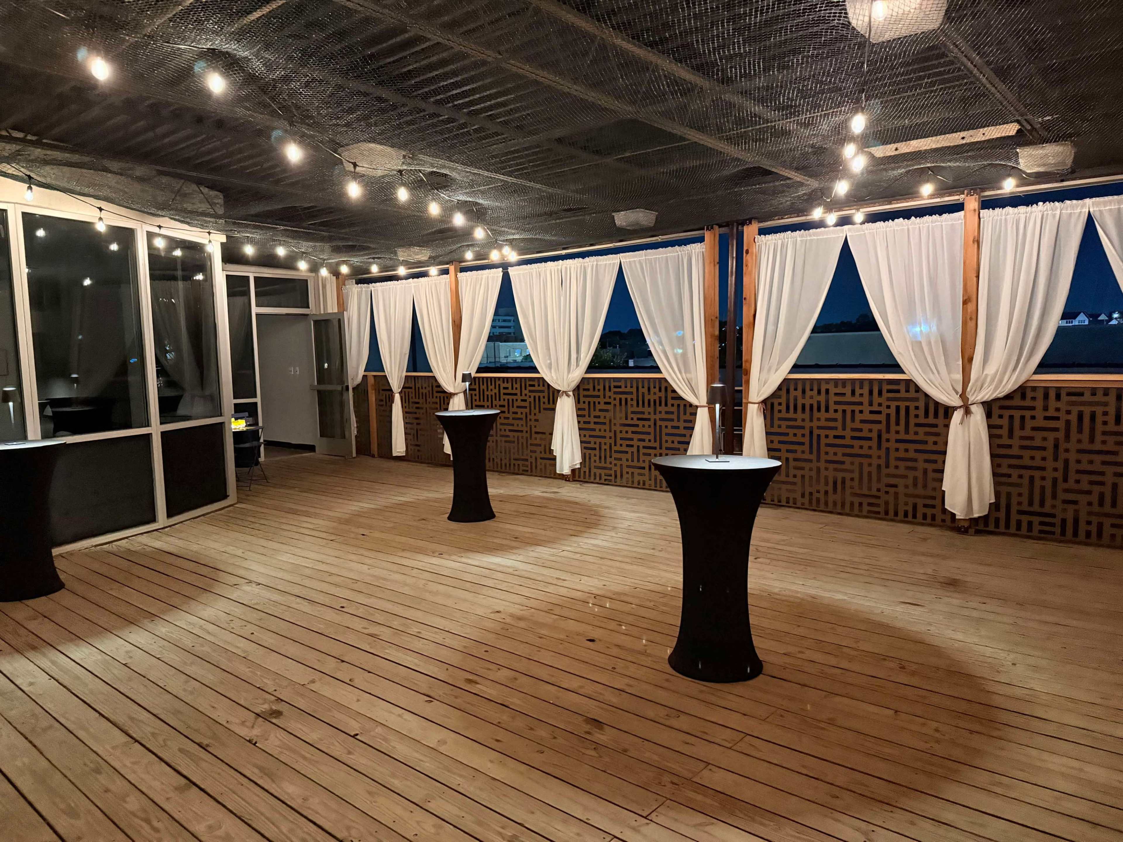 The image shows an empty event space with wooden flooring, featuring several tall tables and sheer curtains draped around the perimeter, illuminated by string lights.