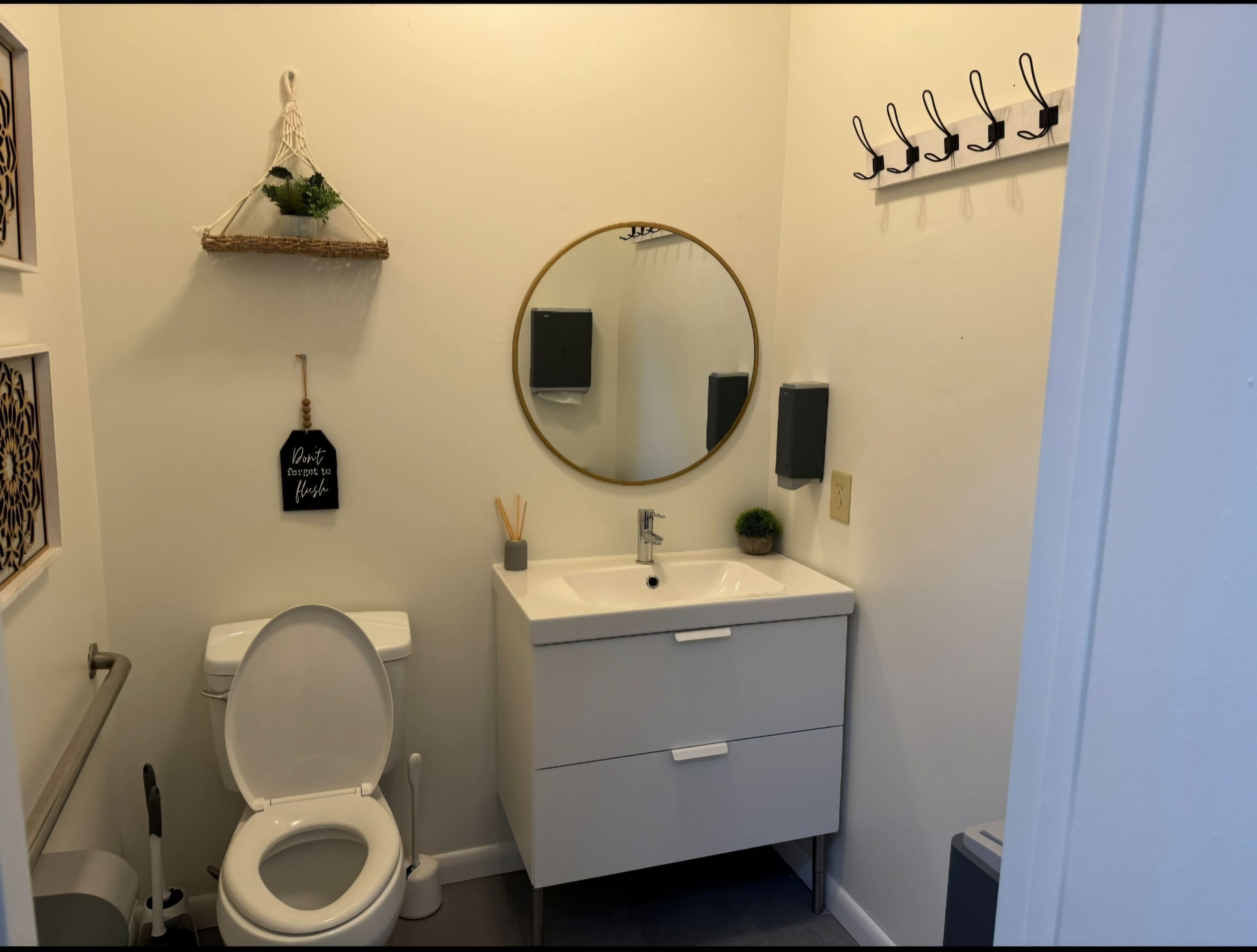 A small bathroom featuring a round mirror above a modern sink and vanity, a toilet, and wall-mounted hooks for hanging items.