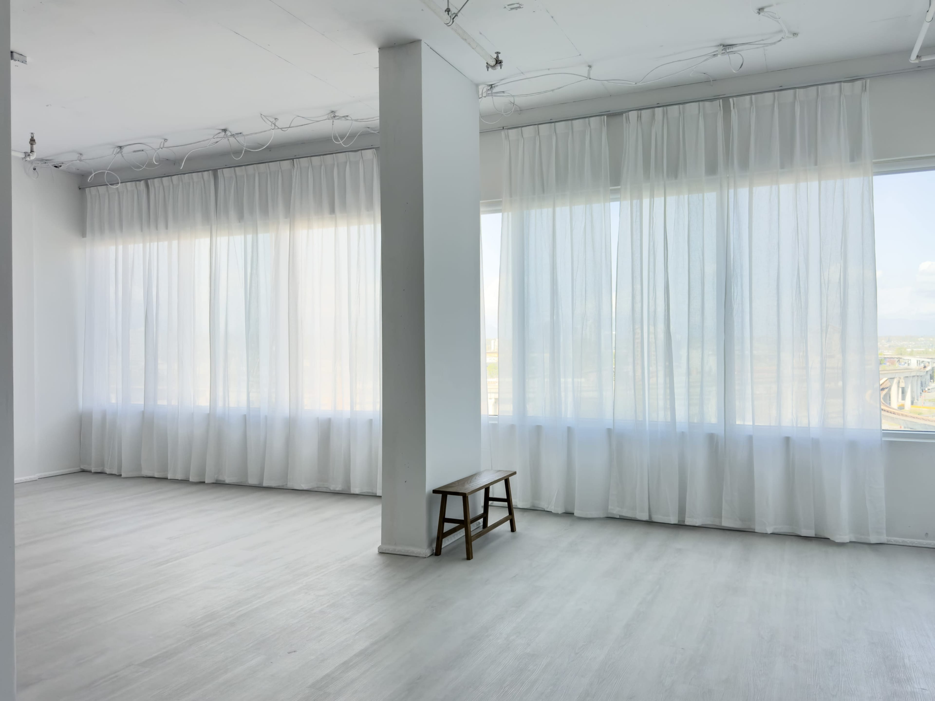 The image shows a bright, empty room with large windows covered by sheer white curtains and a wooden bench placed centrally.