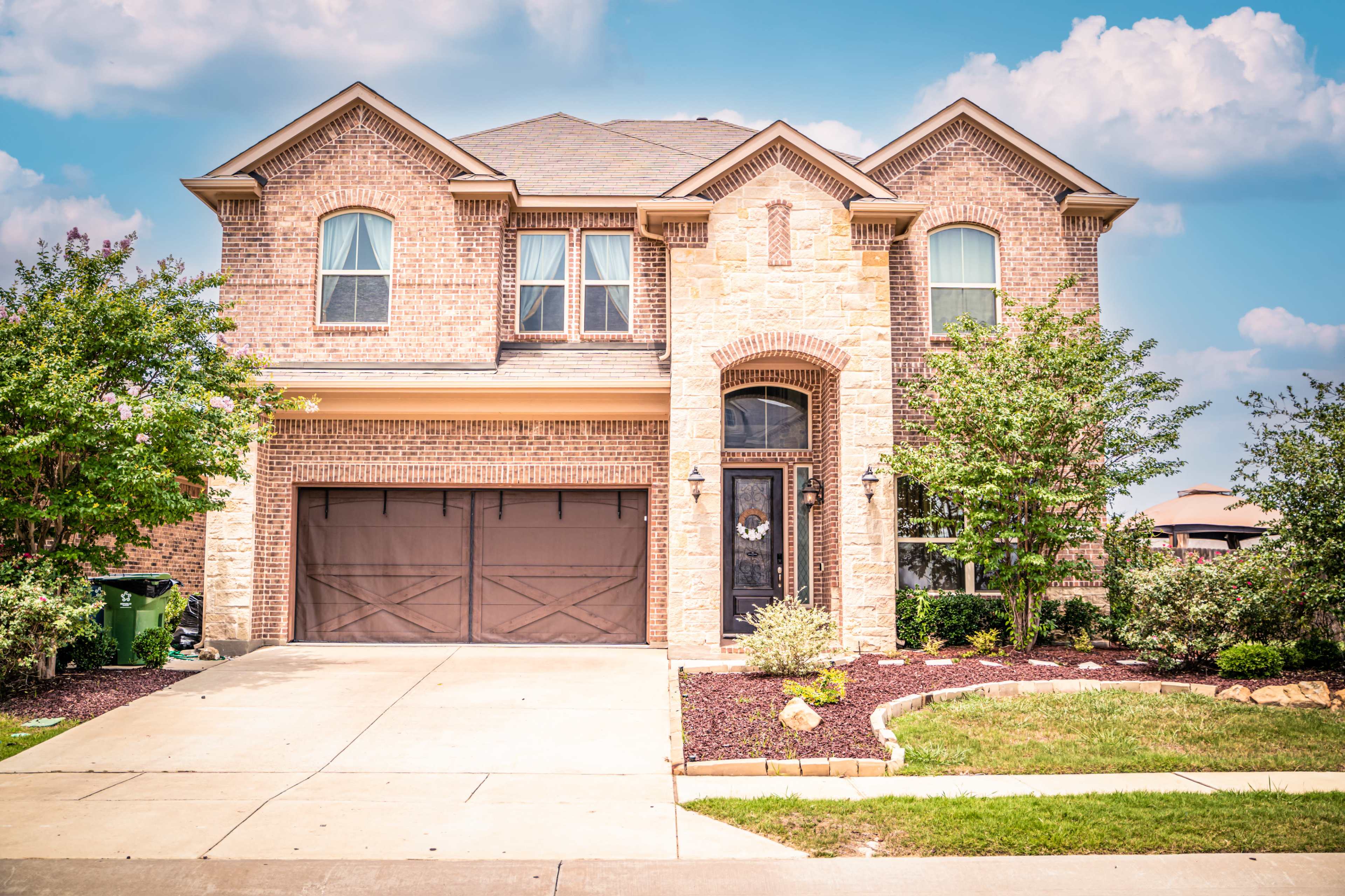 A two-story brick house with a stone accent, a double garage, and landscaped shrubs in the front yard.