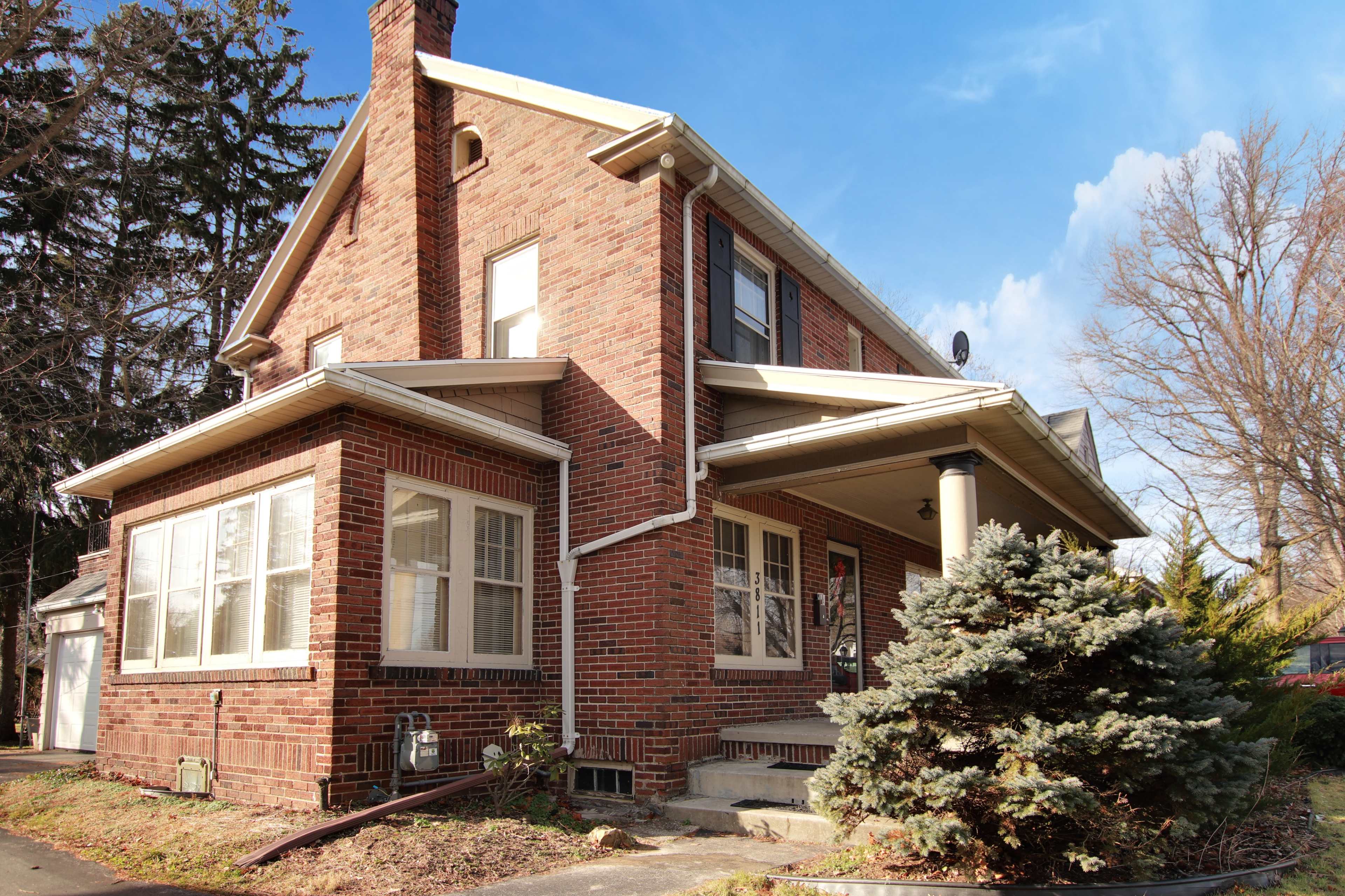 A two-story brick house features a front porch and large windows, surrounded by trees and a landscaped yard.