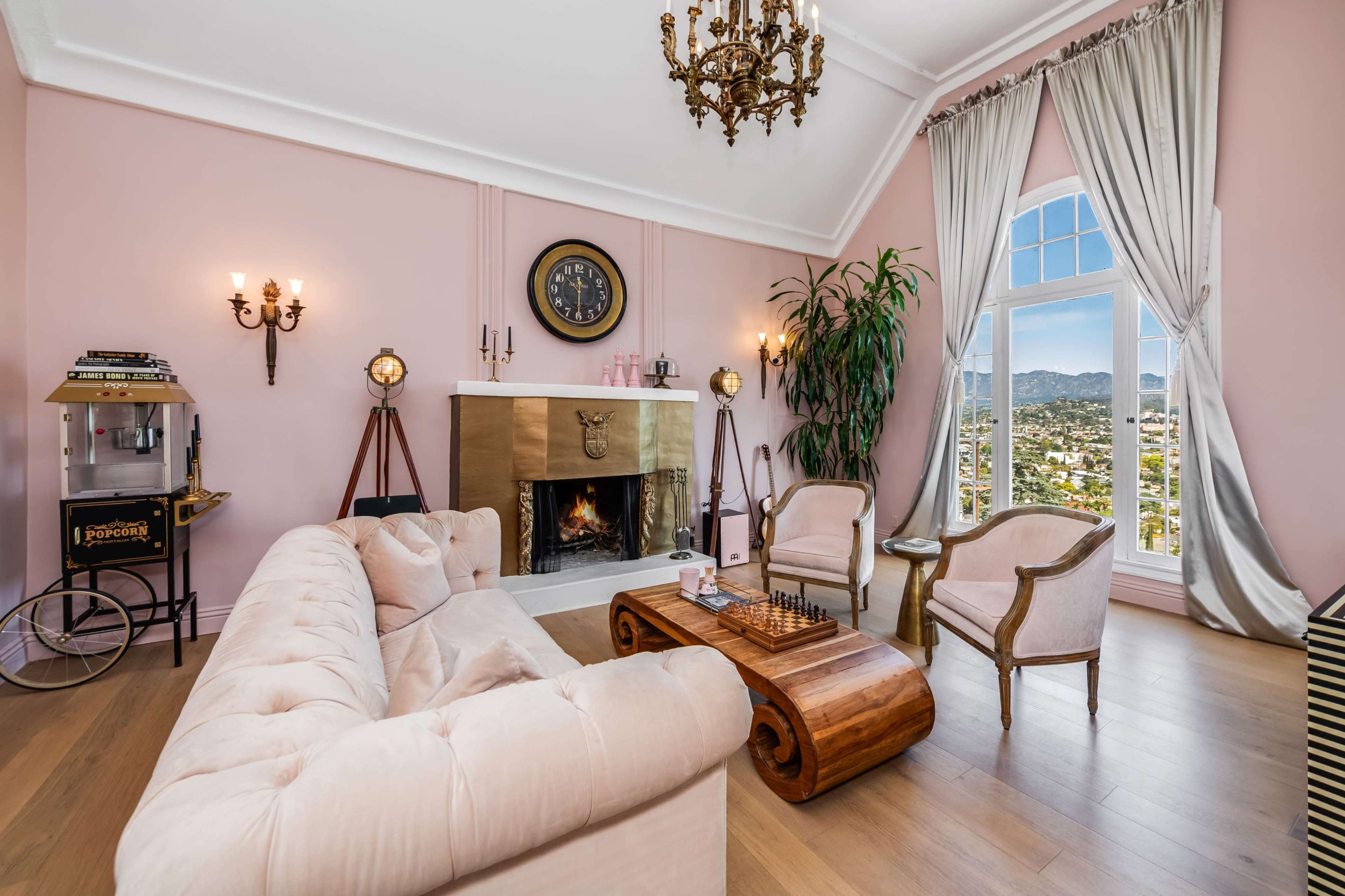 The image shows a stylish living room with a blush pink color scheme, featuring a fireplace, large windows with a mountain view, and elegant furniture arranged around a wooden coffee table.