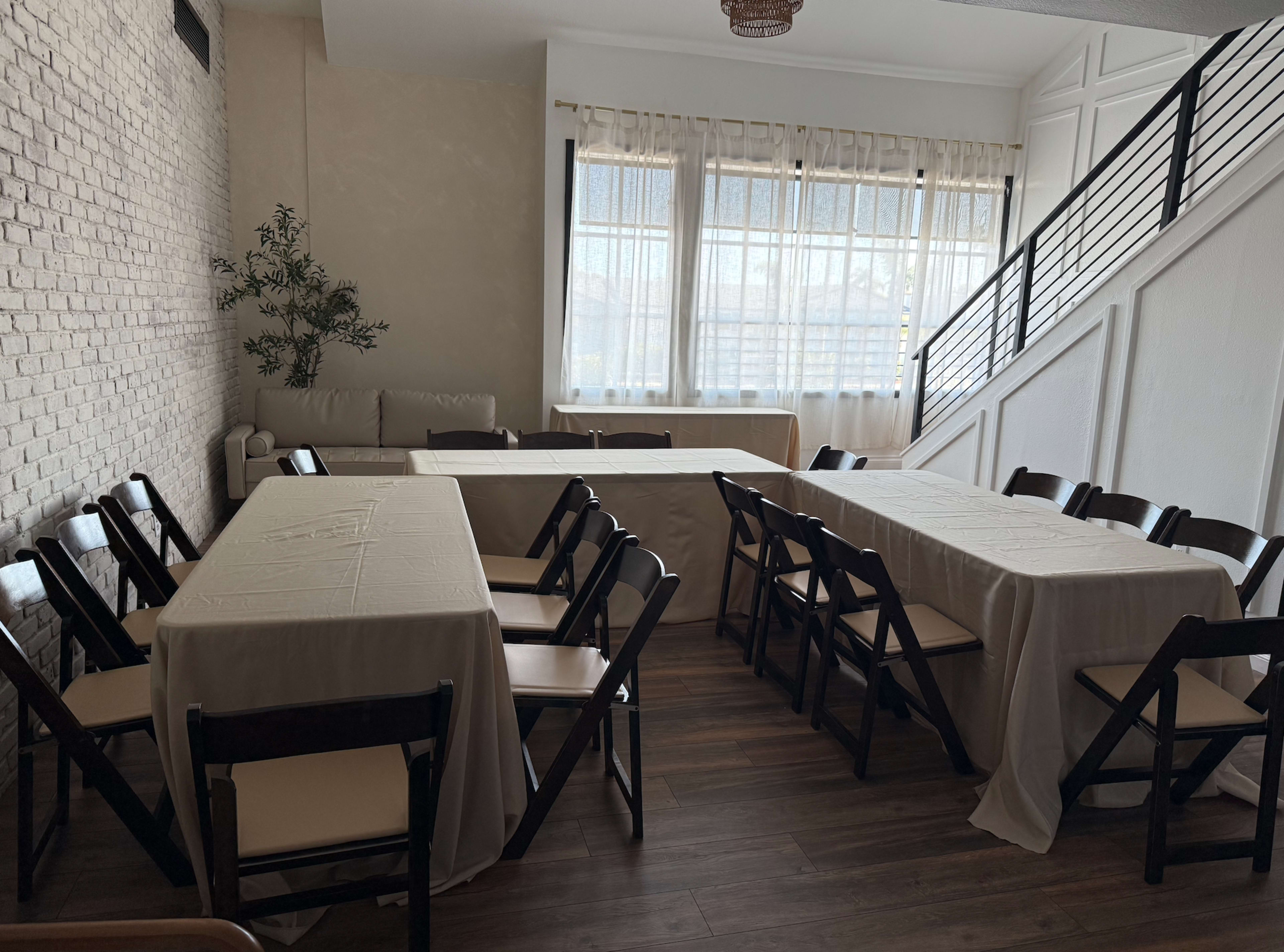The image shows a bright, modern room with several tables covered in white cloth and black folding chairs arranged around them, along with a couch and large windows letting in natural light.