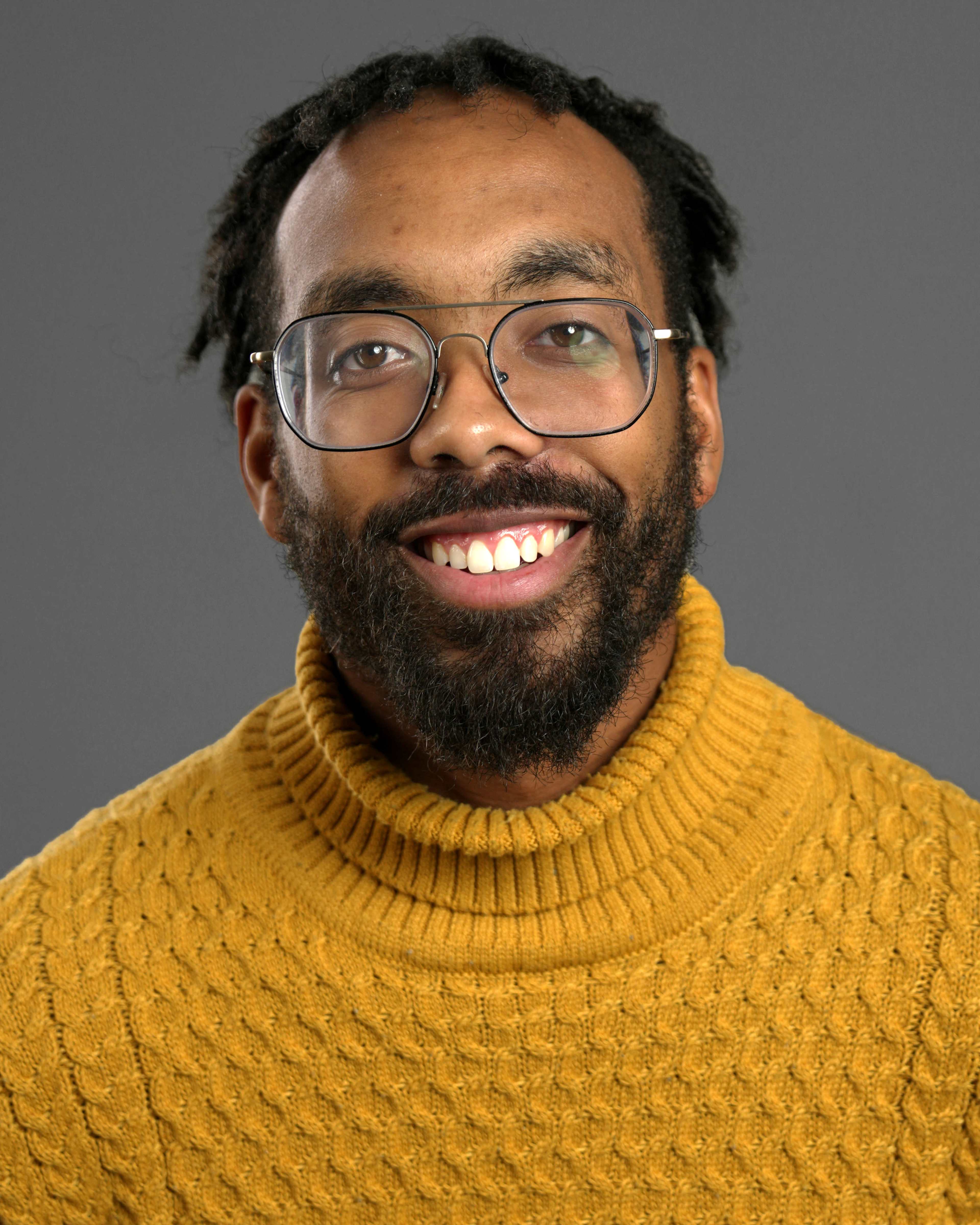 A man with glasses and dreadlocks smiles while wearing a yellow turtleneck sweater against a gray background.