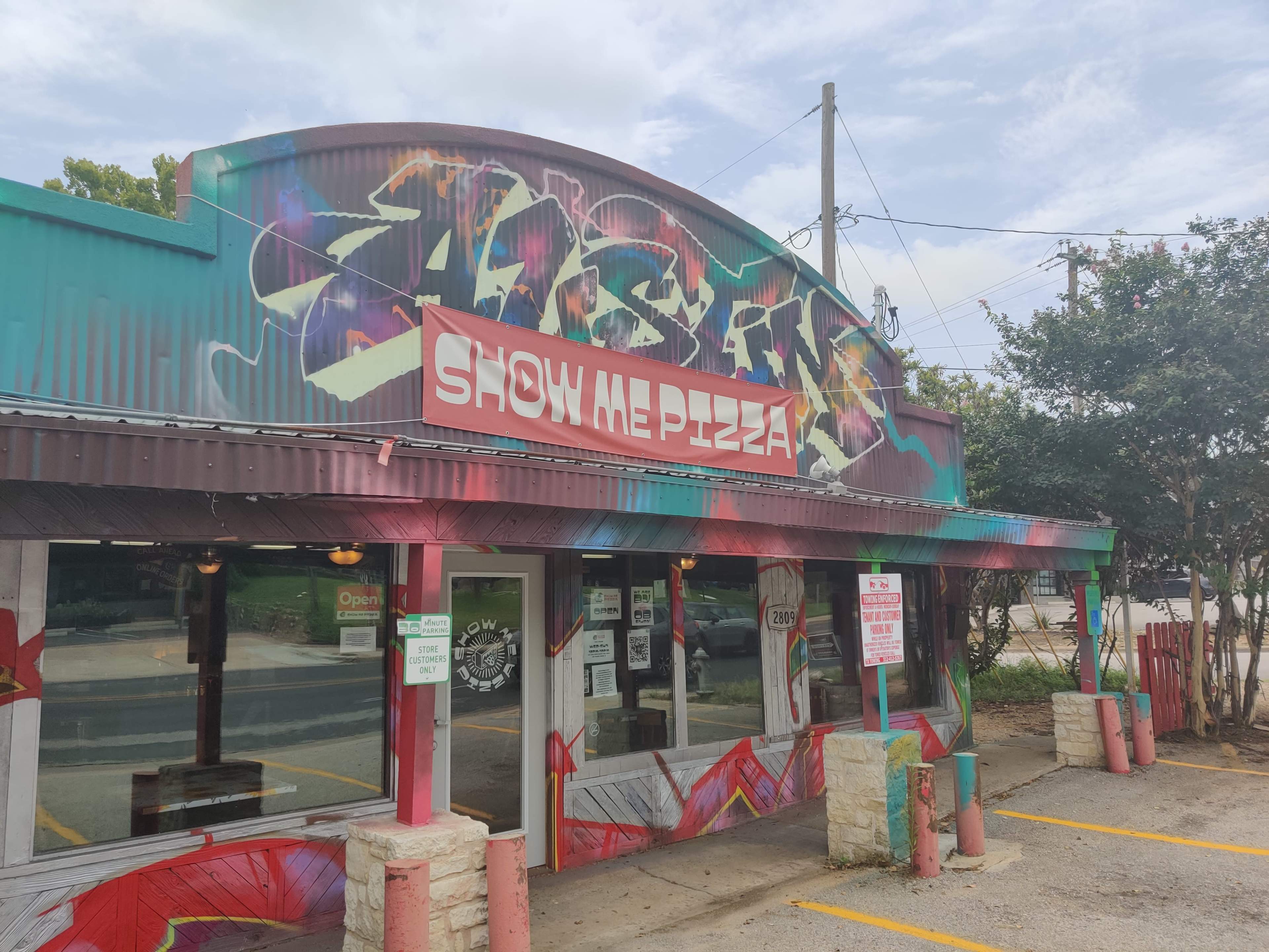 A pizza restaurant with colorful graffiti and a prominent sign reading "SHOW ME PIZZA" features an open entrance and parking spaces in front.