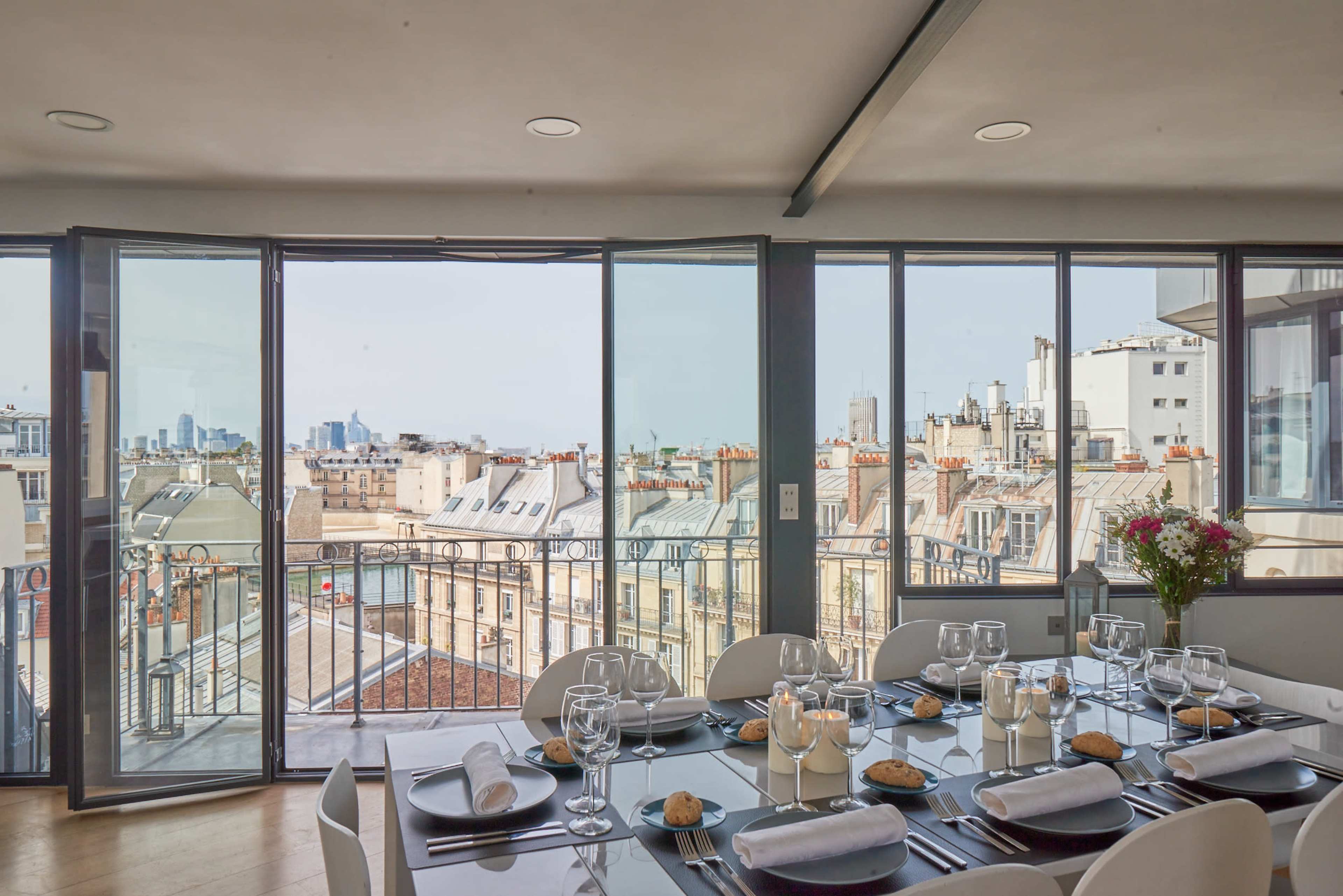 A dining area features a long table set with glassware and cookies, overlooking a cityscape with modern buildings and rooftops through large glass windows.