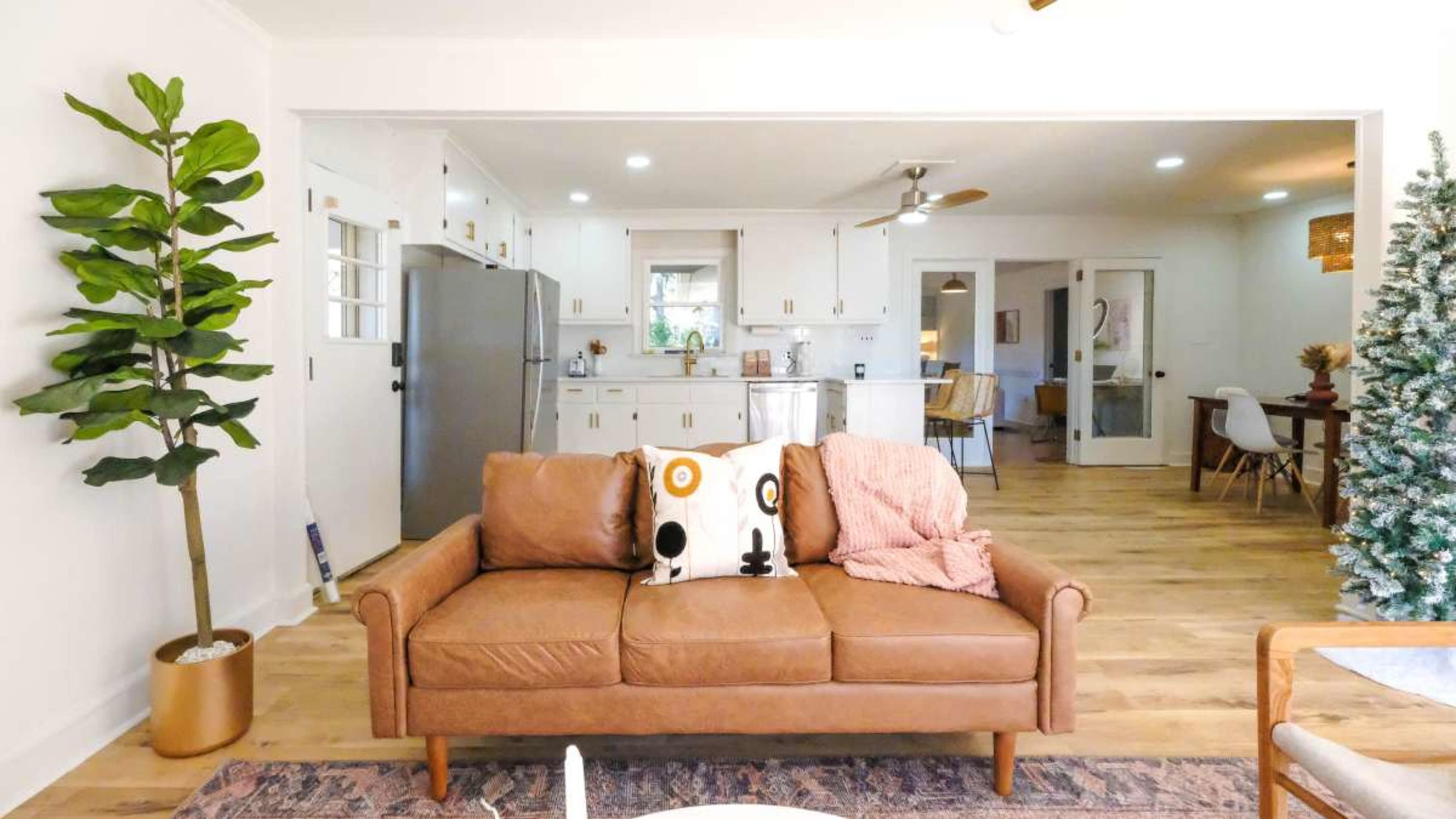 The image shows a cozy living room with a brown leather sofa adorned with decorative pillows and a view of an open kitchen in the background.