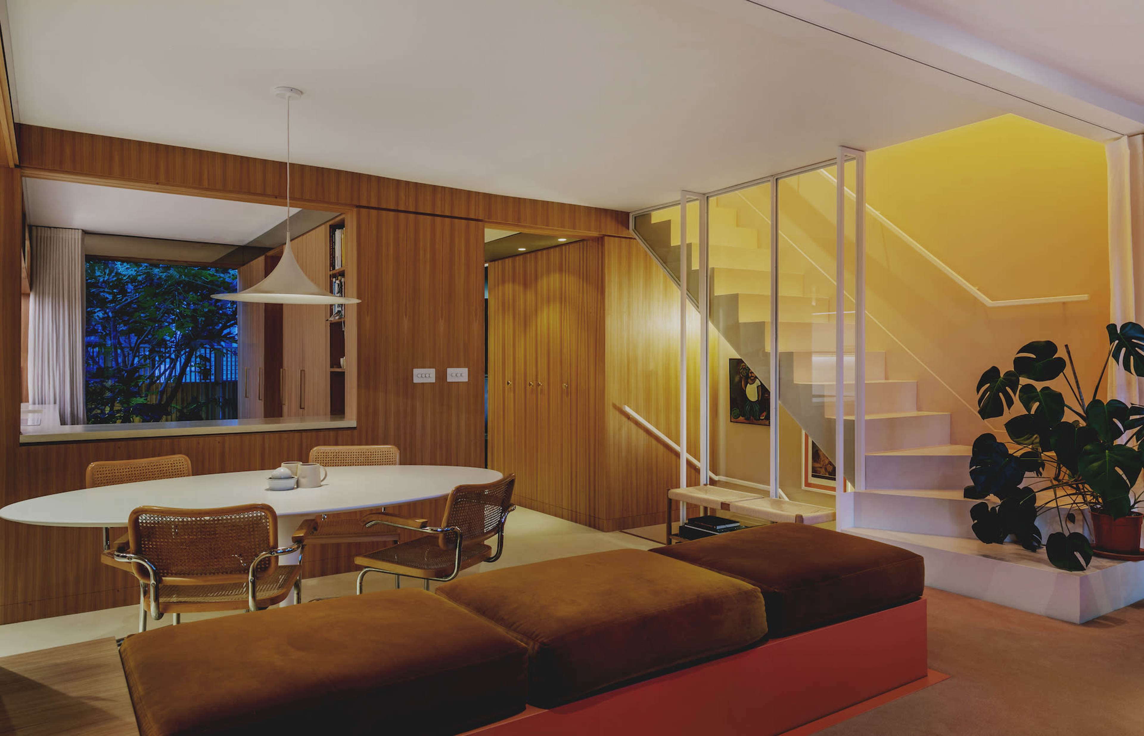 A modern living area features a circular dining table, a sectional couch, and a staircase leading to an upper level, with warm lighting illuminating wooden walls and greenery visible through a window.