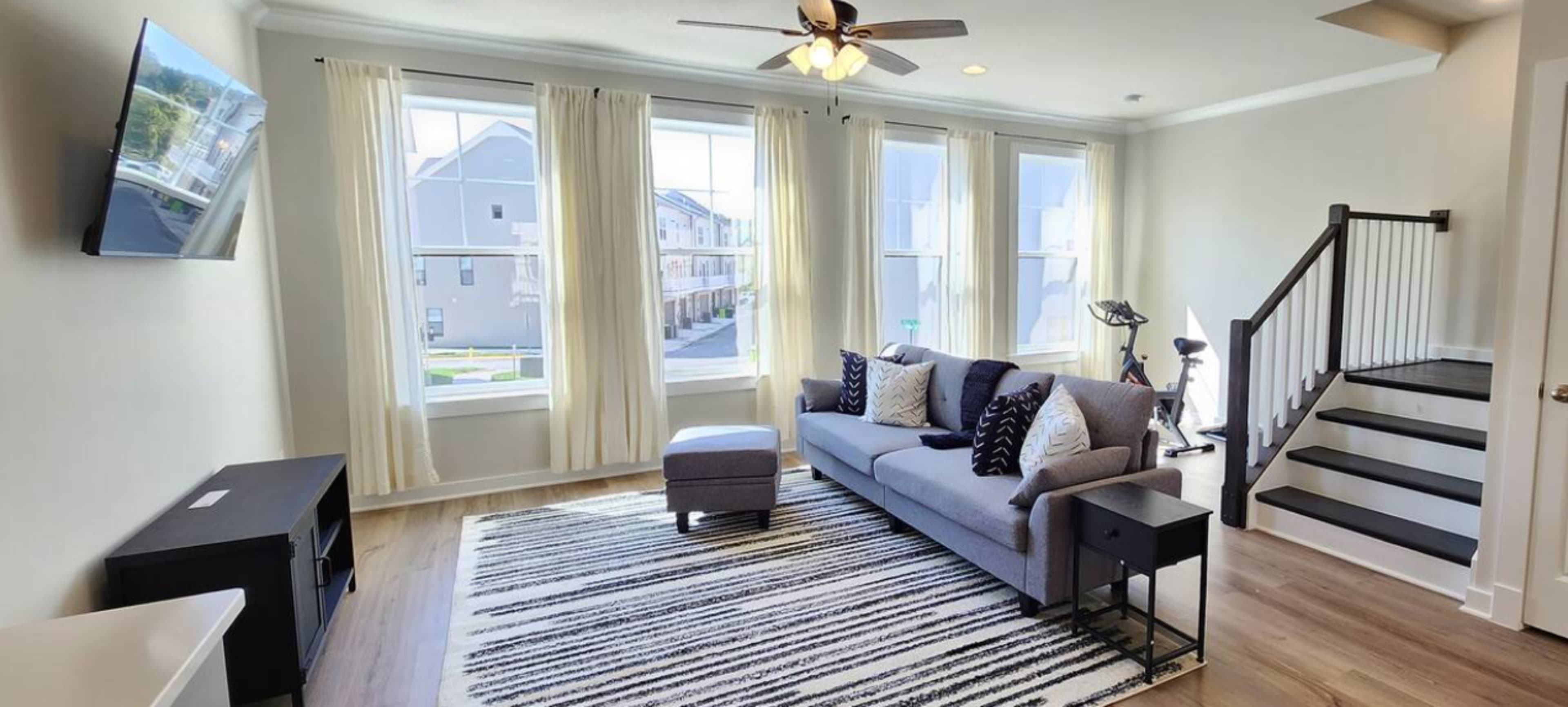 A living room with large windows, a gray sofa with patterned pillows, a small side table, and a black TV mounted on the wall, with stairs leading up to another level.