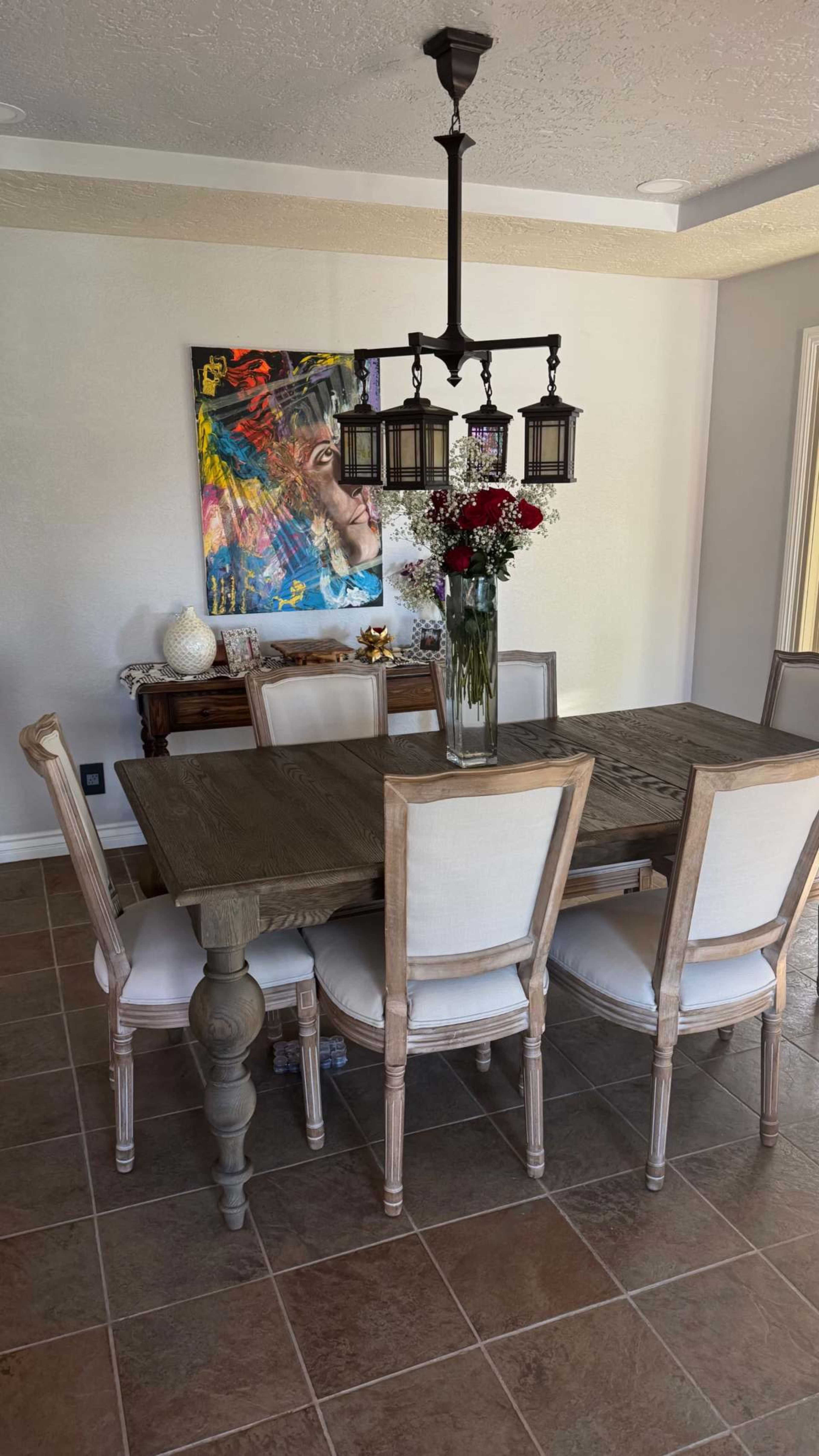 The image shows a dining room with a wooden table surrounded by six chairs, a large flower vase in the center, and a colorful painting on the wall.