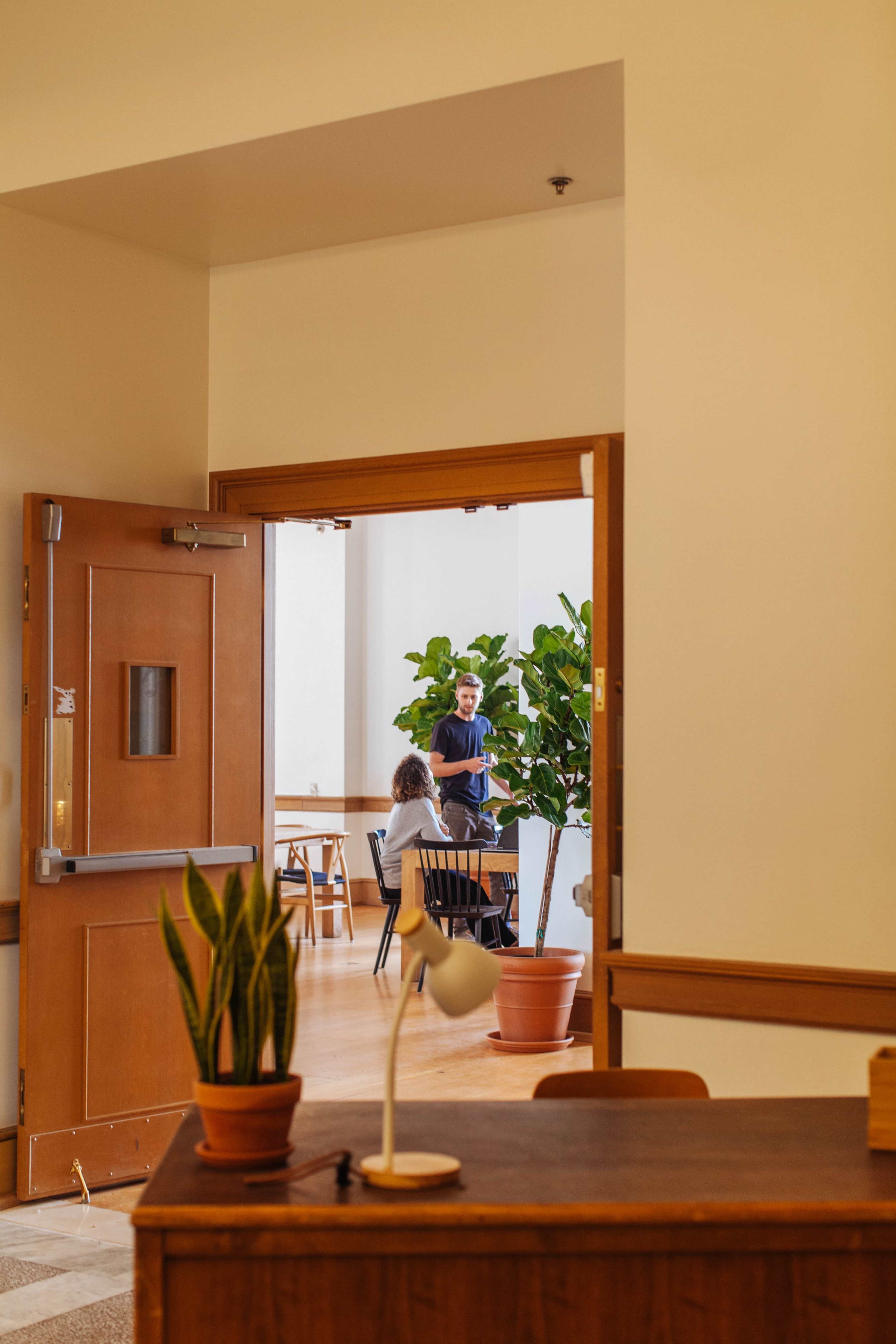 A person stands conversing with another individual seated at a table in a well-lit room, framed by an open doorway with a plant in the foreground.