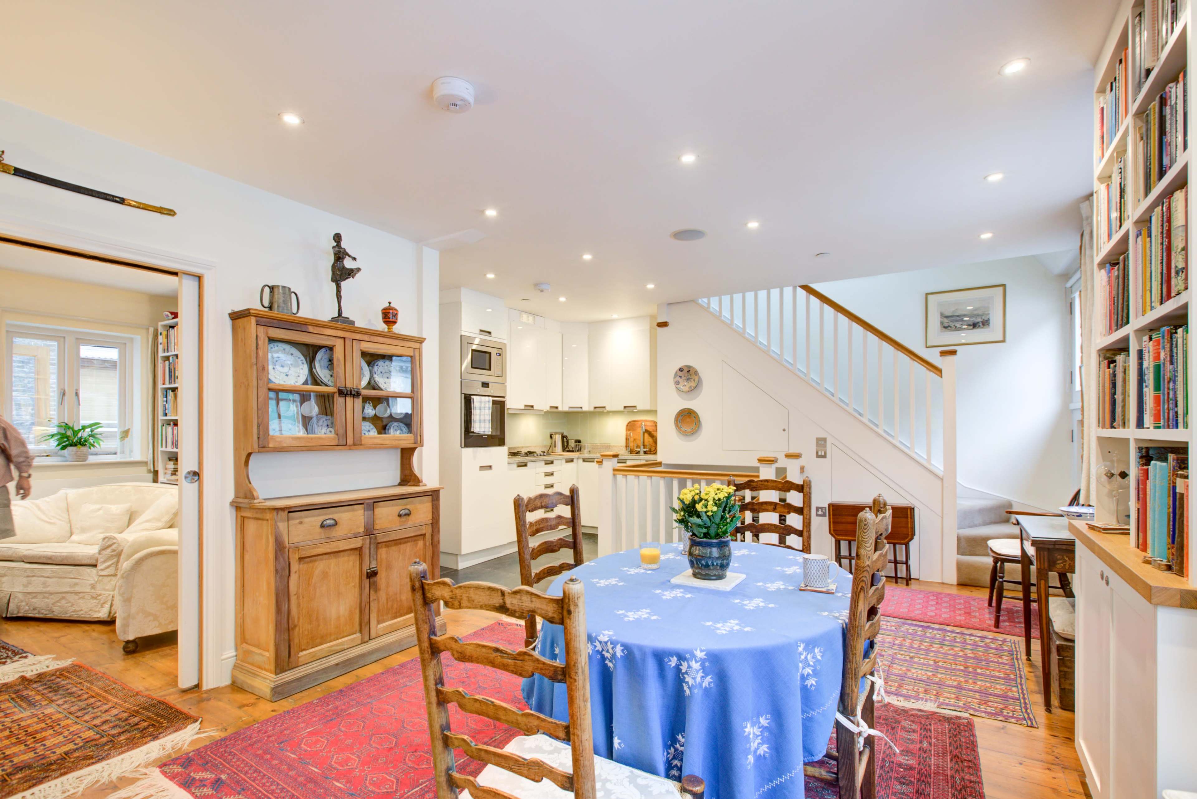 A cozy dining area with a blue tablecloth-covered table surrounded by wooden chairs, leading into a well-lit kitchen and living space.