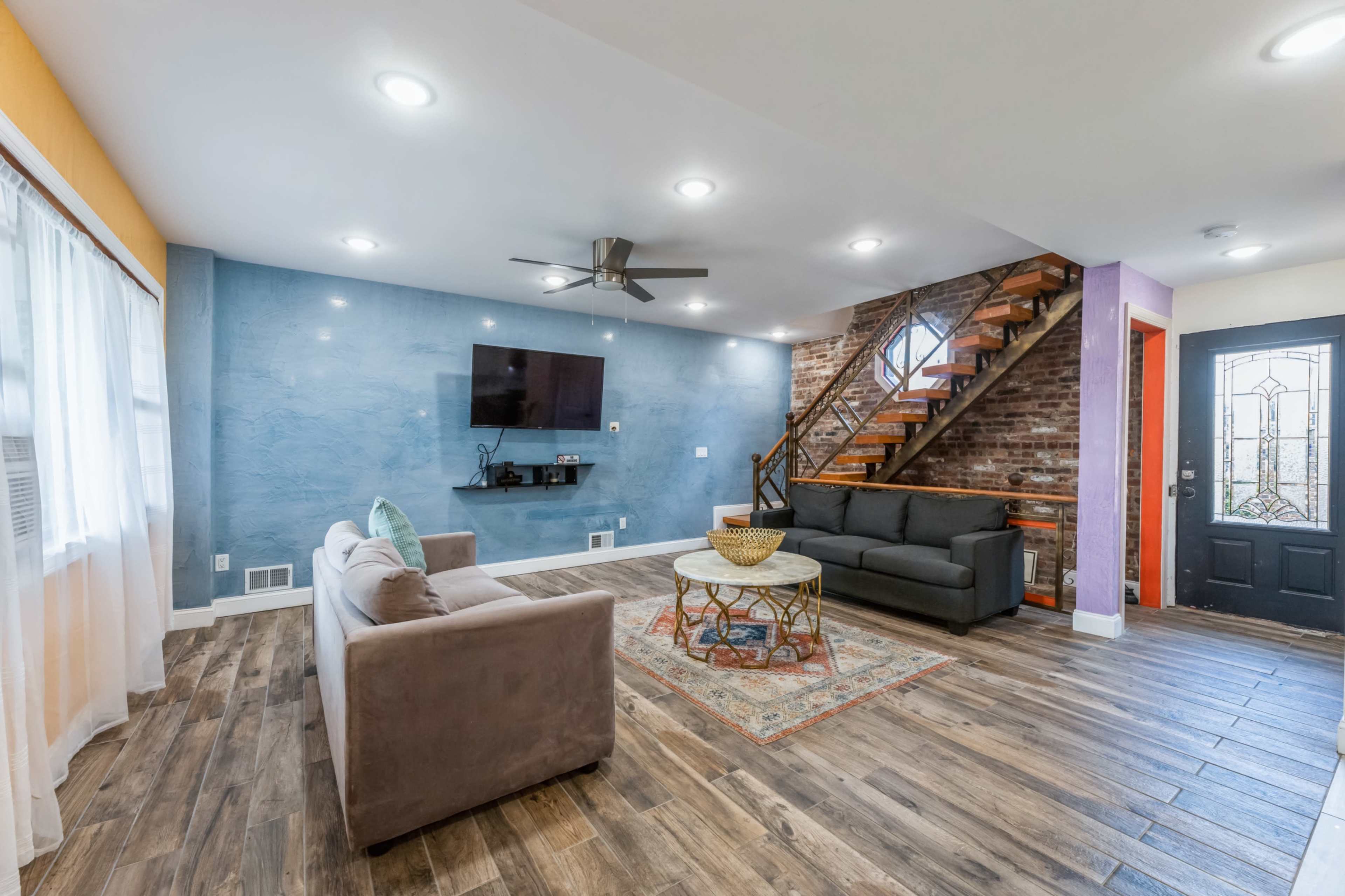 A living room with a blue accent wall, two sofas, a round coffee table, a television mounted on the wall, and a staircase leading to an upper level.