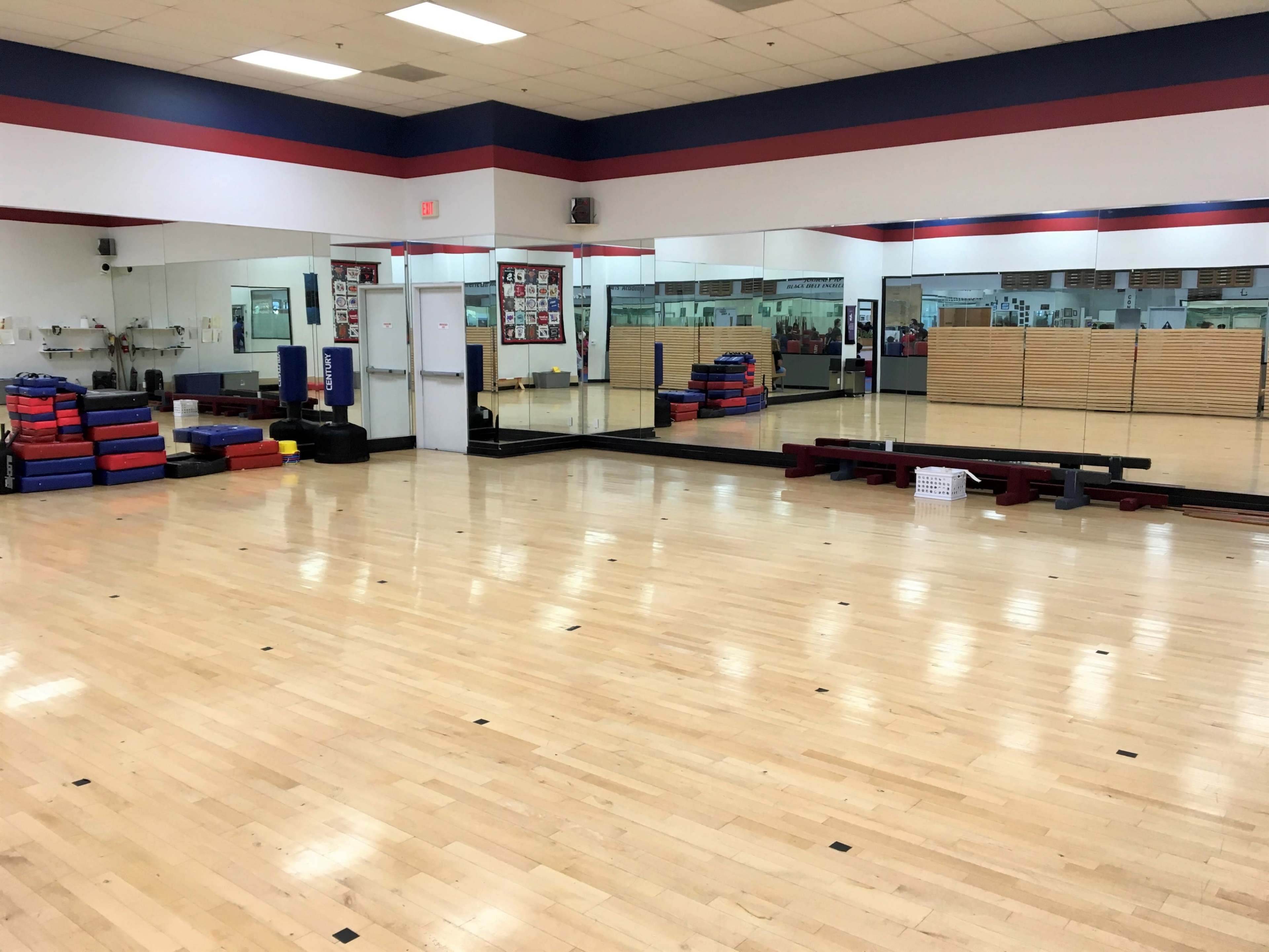 An empty gymnasium with wooden flooring, large mirrors on one wall, colorful gym mats stacked in one corner, and various fitness equipment scattered around.