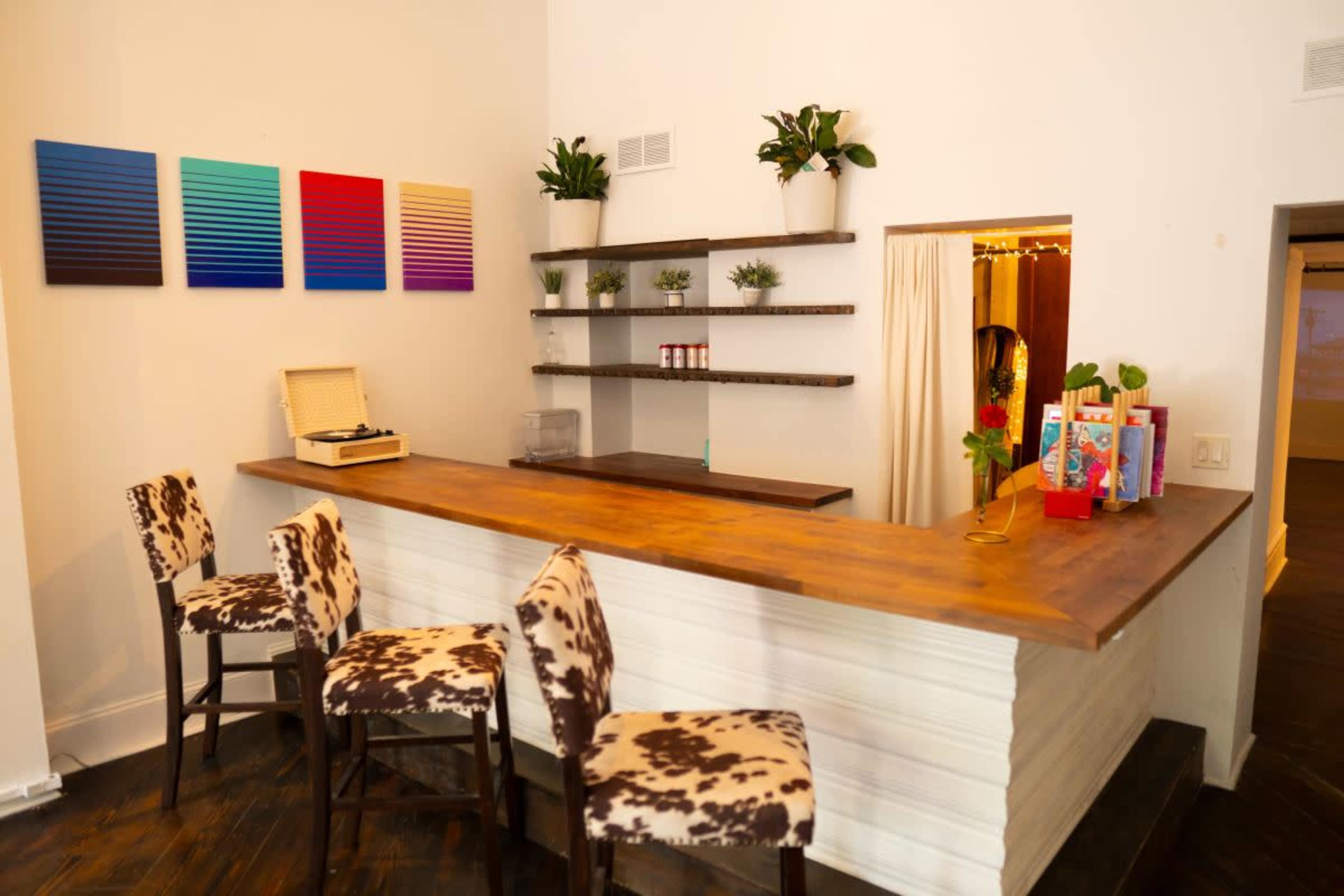A modern bar area features a wooden counter with high stools, shelves displaying small plants, and colorful artwork on the wall.