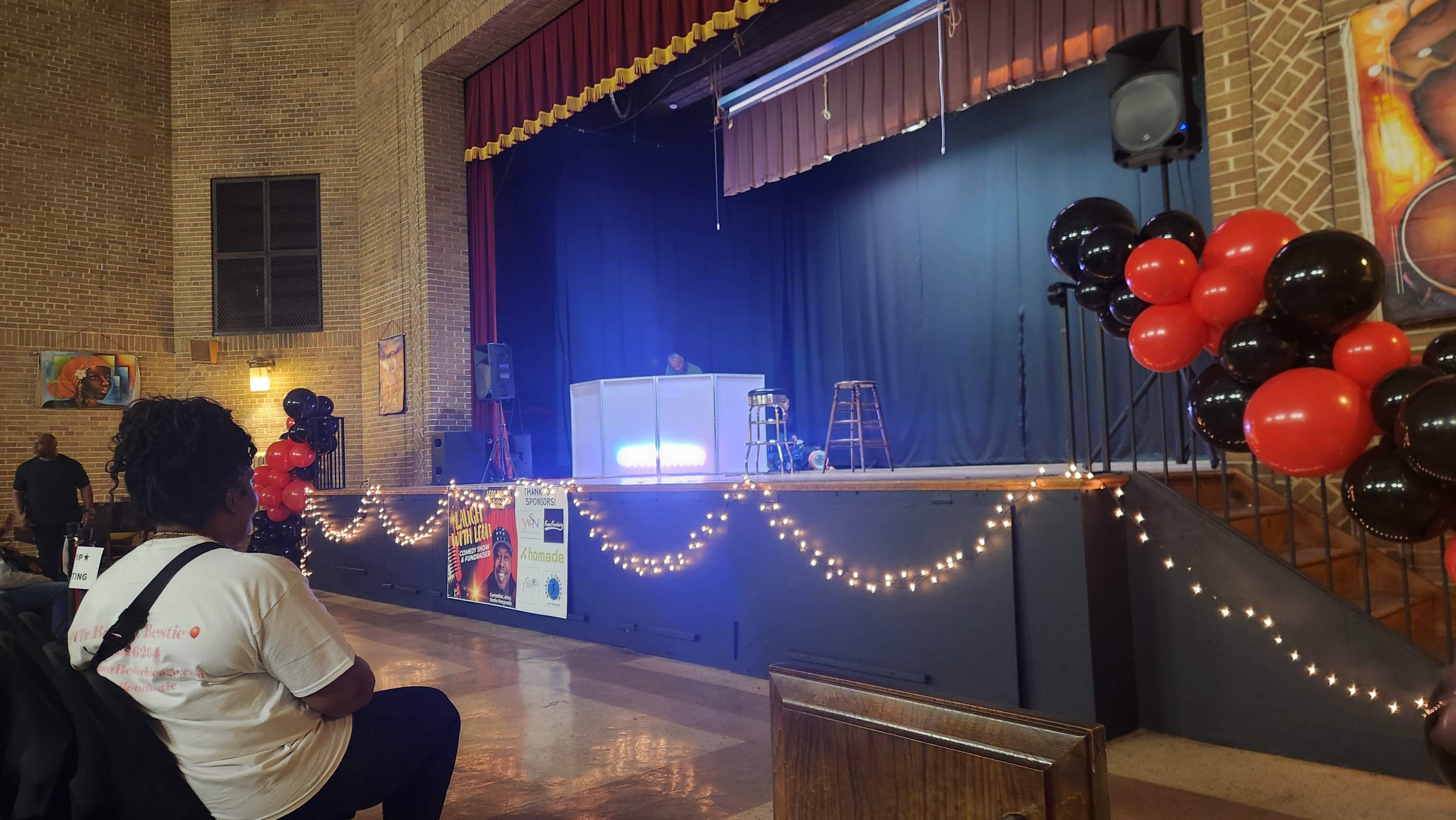 A stage is set with a black curtain and lighting, surrounded by decorations of red and black balloons, while a person sits in the audience facing the stage.