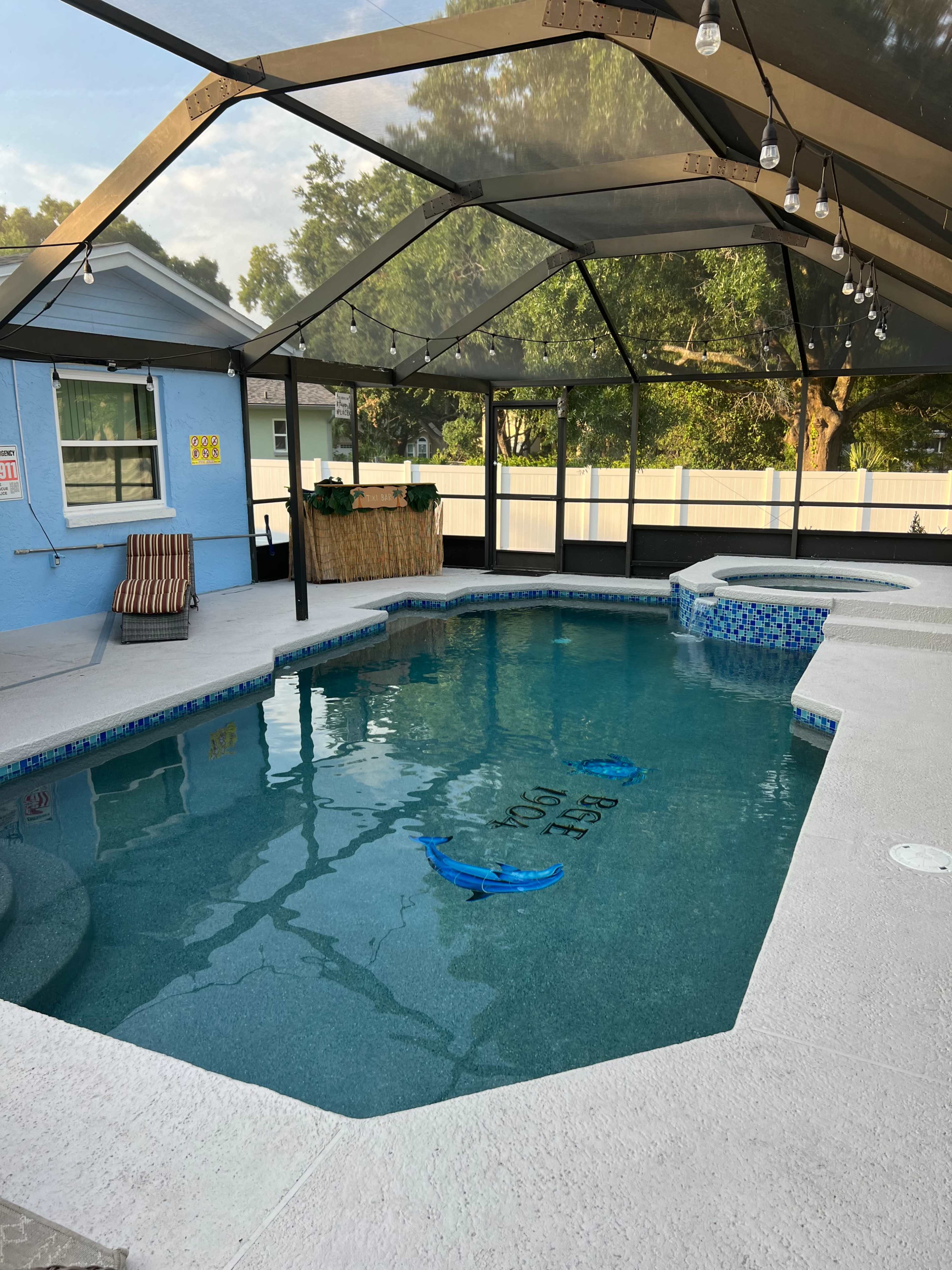A screened-in swimming pool area with a blue pool, a lounge chair, and a small bar under a canopy of string lights.