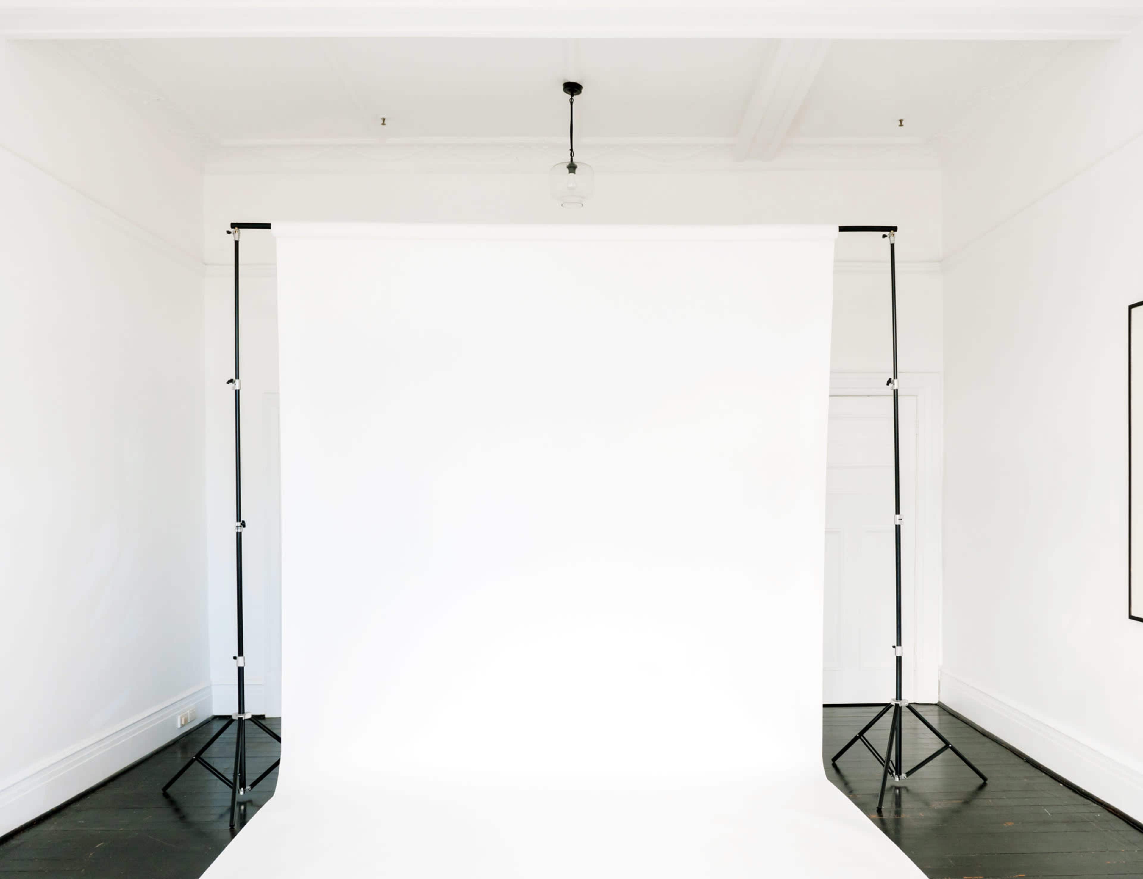 A photography studio with a blank white backdrop suspended between two black stands in a room with white walls and wooden flooring.