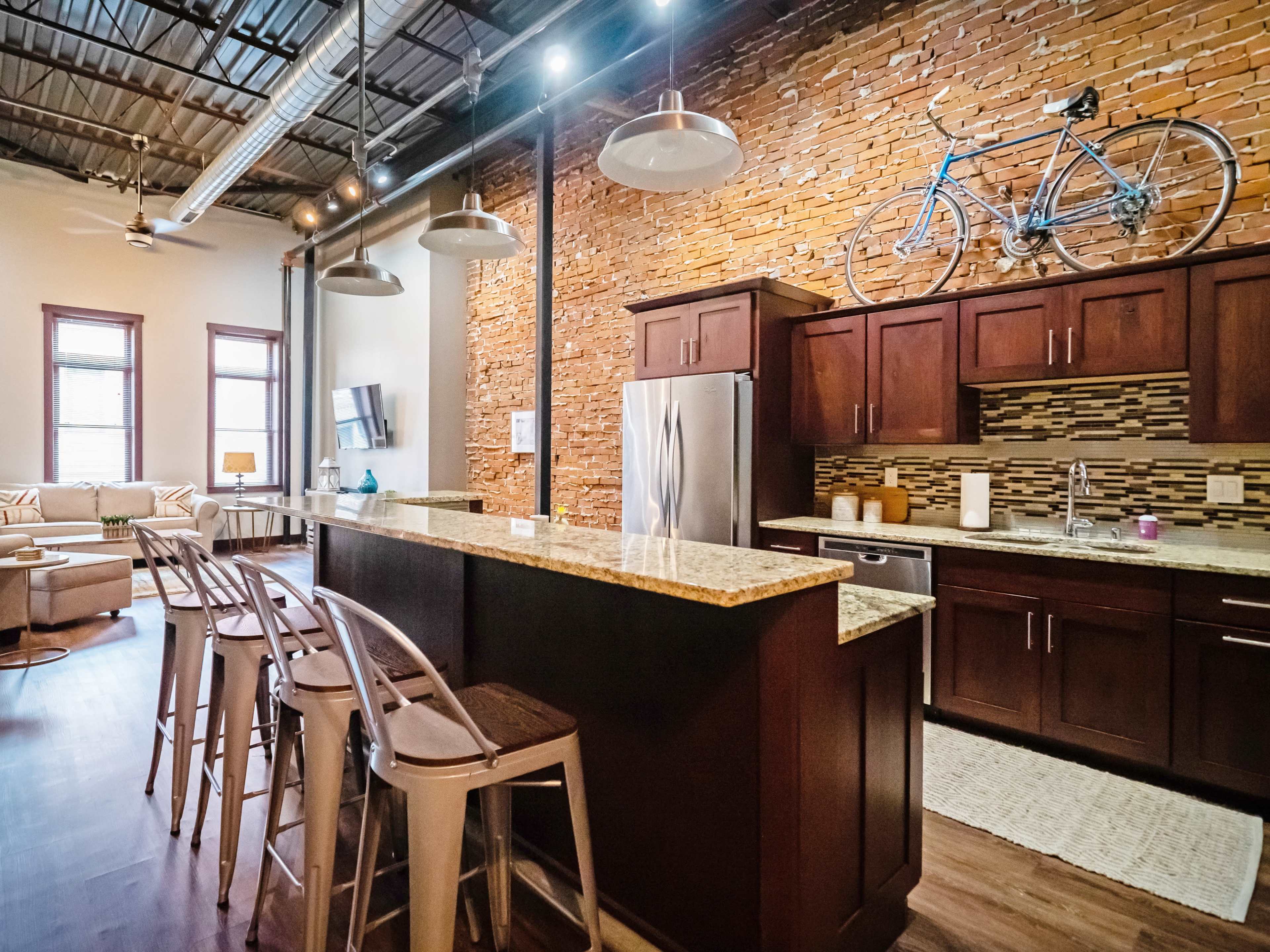 The image depicts a modern kitchen with granite countertops and dark wooden cabinetry, accented by a brick wall and a bicycle mounted above the cabinets.
