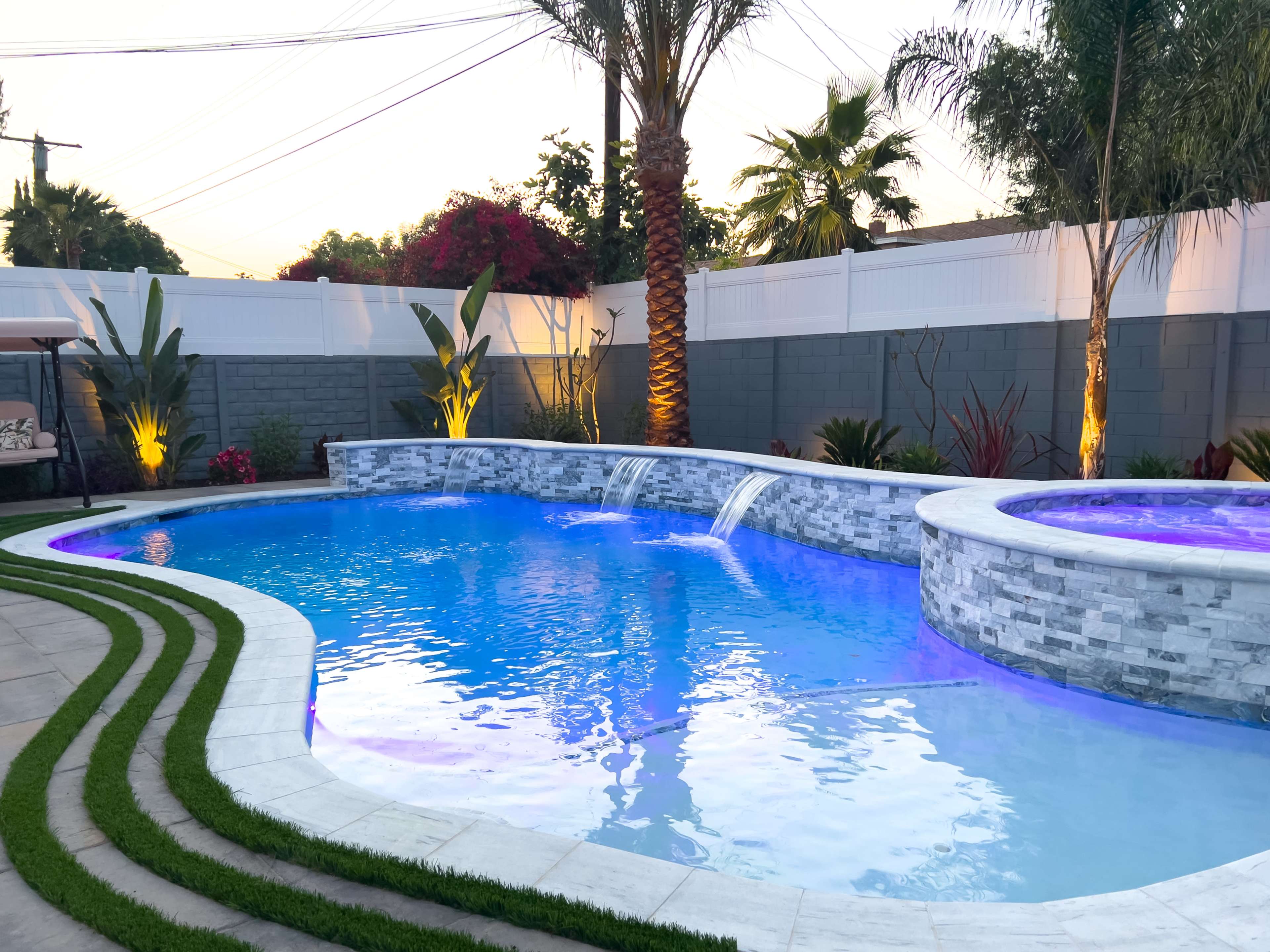 A backyard swimming pool featuring a stone waterfall and a spa, surrounded by landscaped greenery and illuminated at dusk.