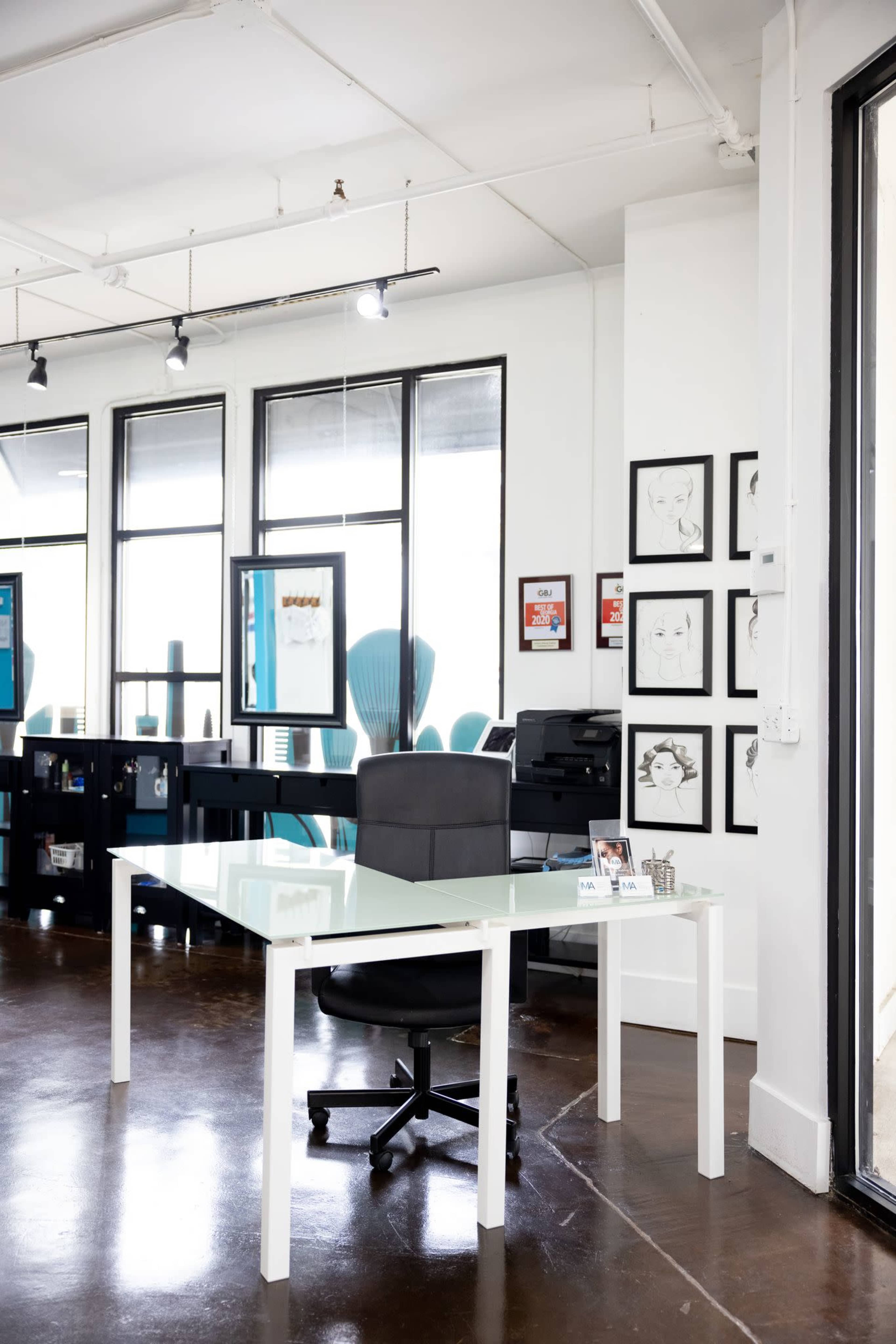 A modern office space features a glass-top desk with a black swivel chair, surrounded by large windows and framed artwork on the walls.