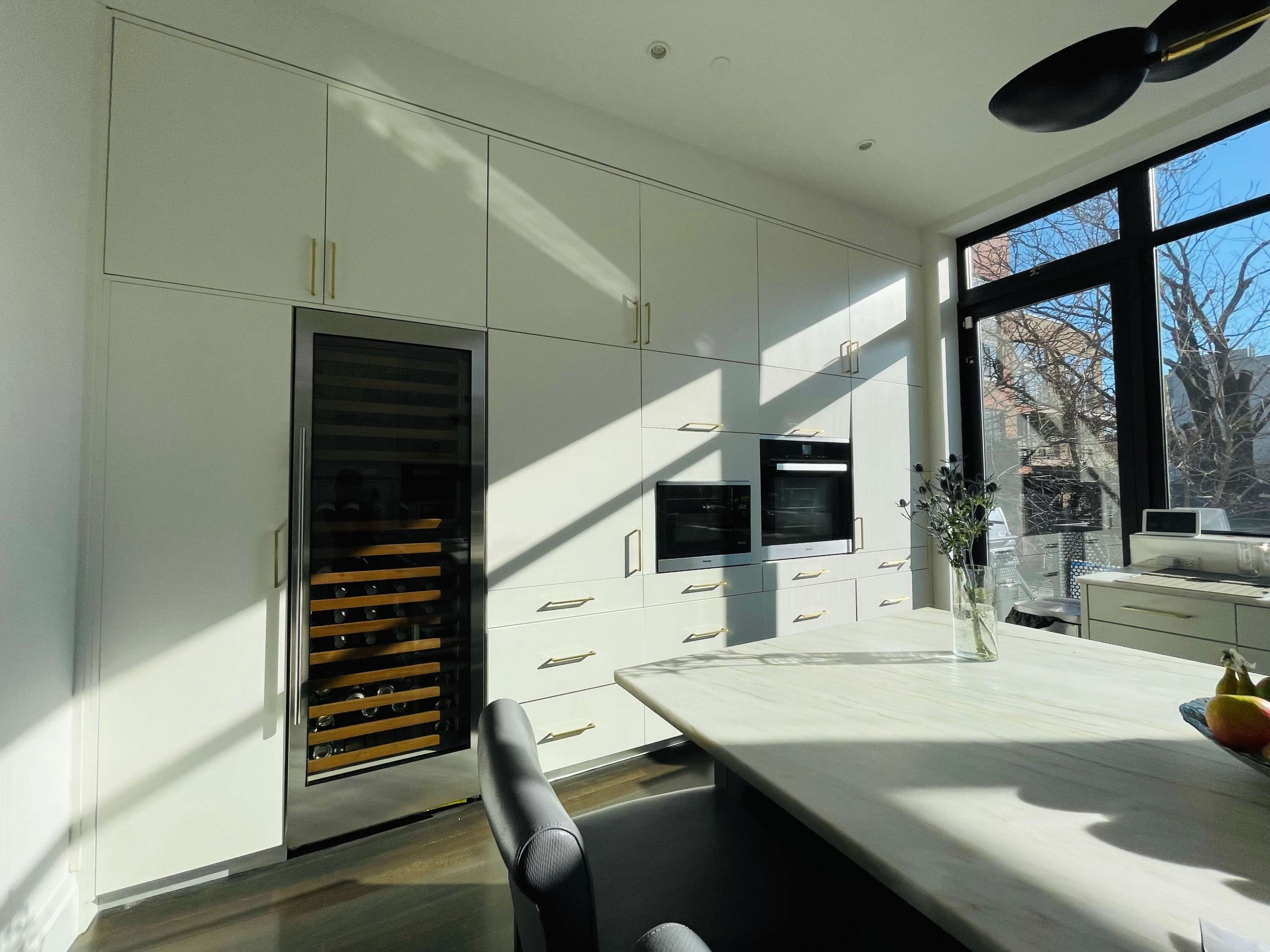 A modern kitchen featuring white cabinetry, a wine cooler, built-in ovens, and a large marble-topped island bathed in natural light.