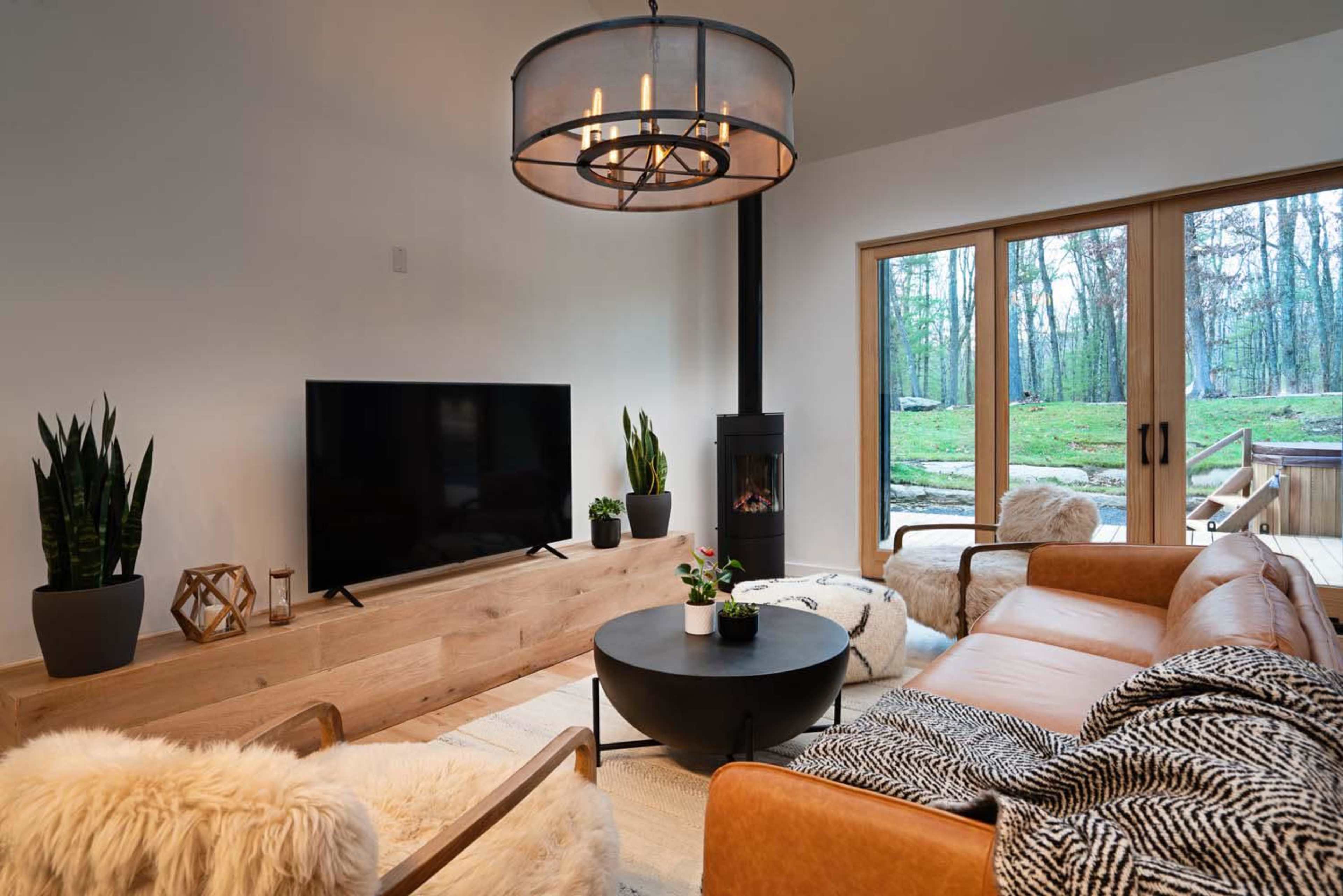 A modern living room features a circular chandelier, a black television on a wooden console, a cozy leather sofa, and large windows overlooking a wooded area.