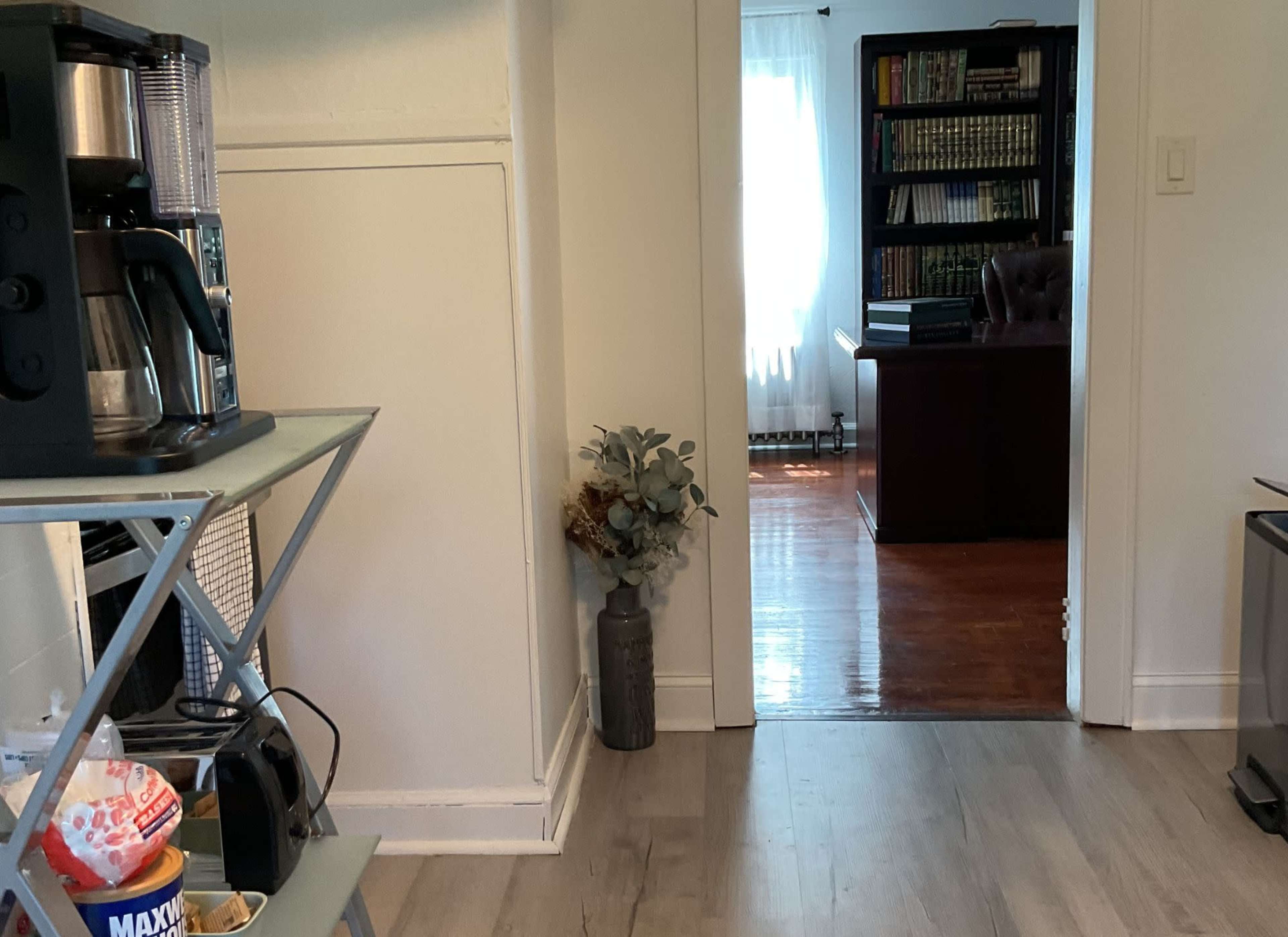 A coffee station is set up on a metal shelf next to a doorway leading to a room with bookshelves and a desk.