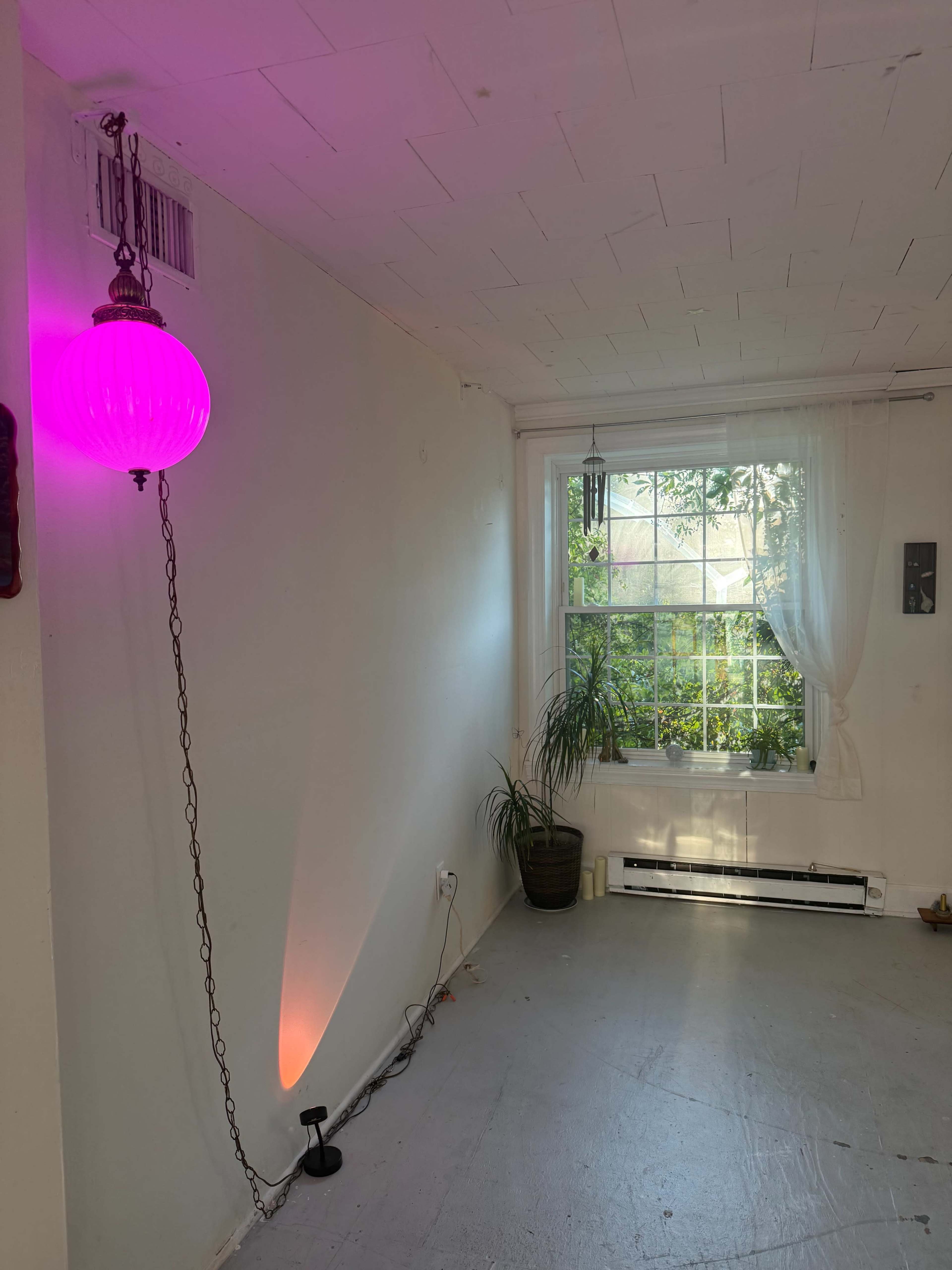 The image shows a room with a pink pendant light hanging from the ceiling, a window with sheer curtains, and a potted plant near the wall.