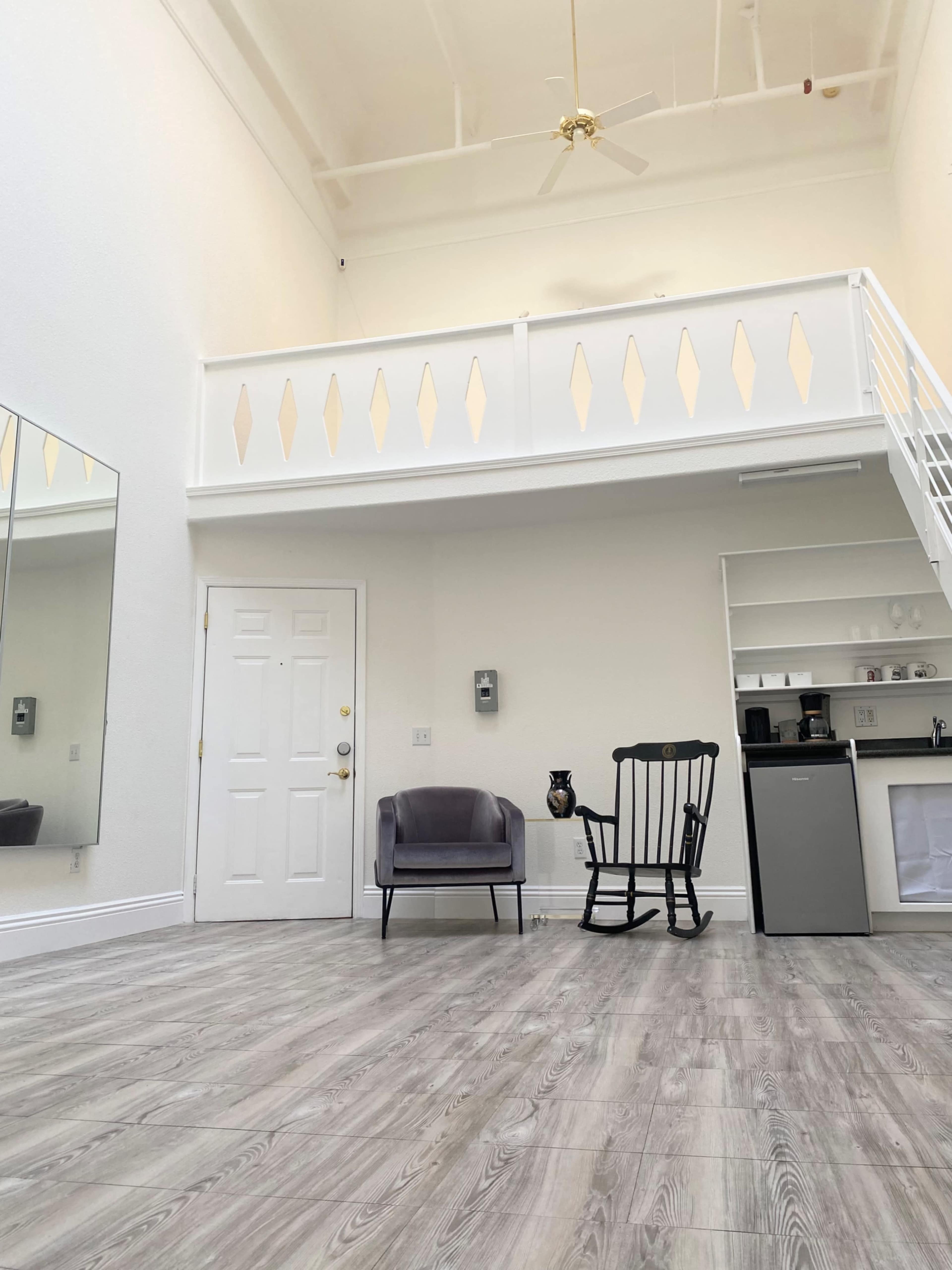 A spacious room with a high ceiling, a rocking chair, a small refrigerator, and a door leading outside, along with a mirrored wall and a mezzanine balcony.