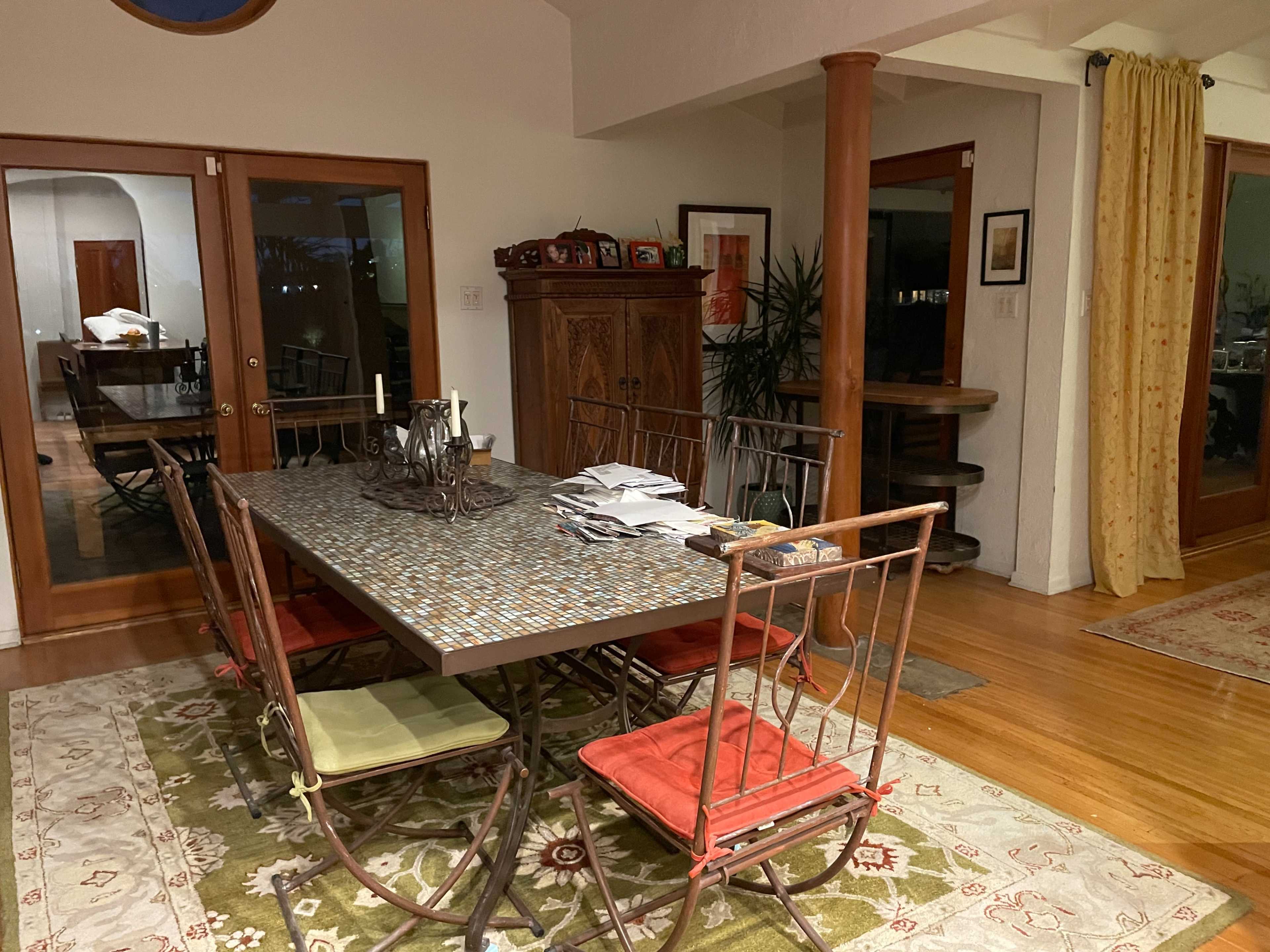 A dining area with a mosaic-topped table surrounded by several chairs, hardwood floors, a plant, and wooden cabinets in a well-lit space.