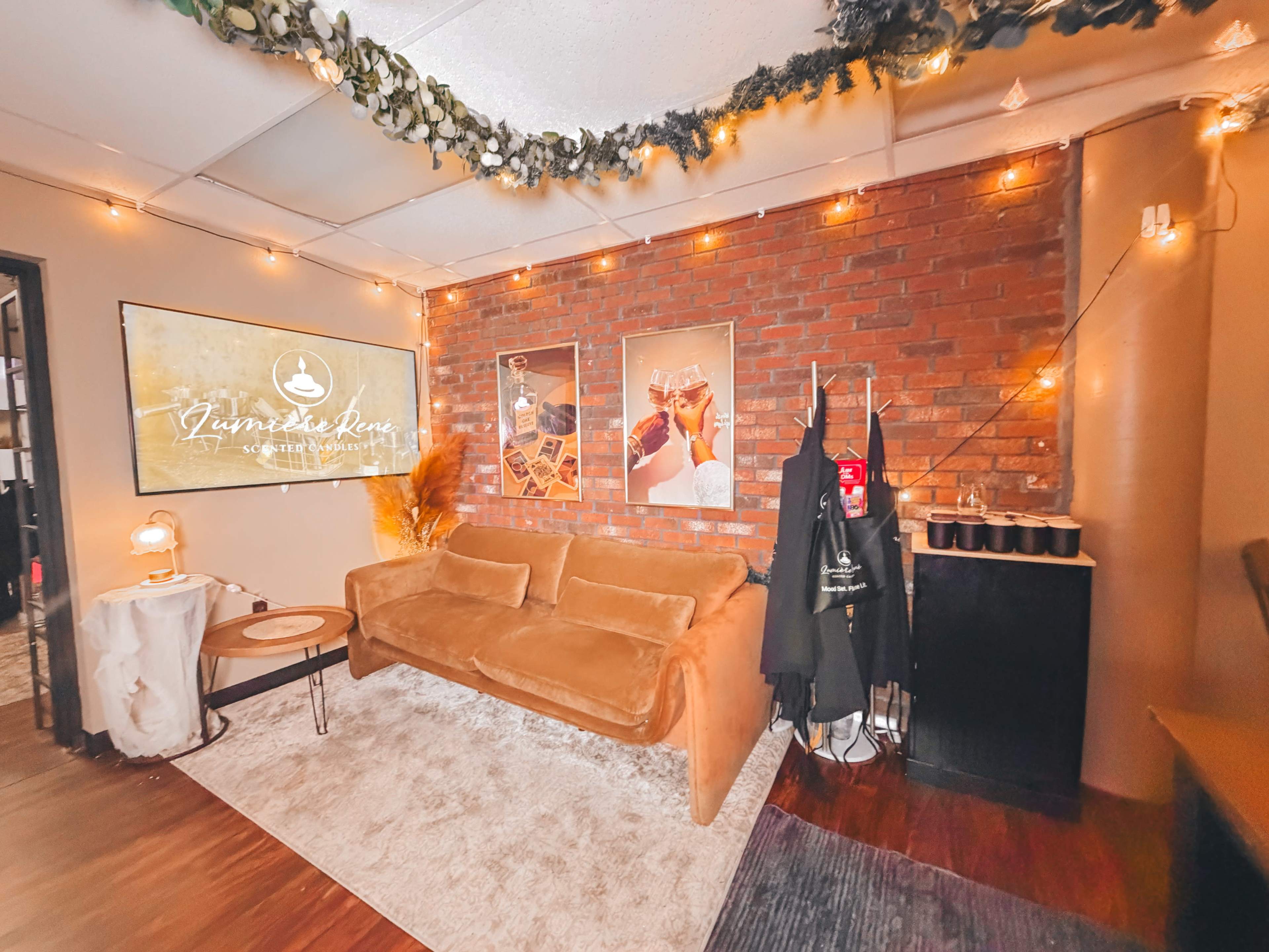 The image shows a cozy lounge area with a brown couch, decorative lighting, art on the walls, and a small table.