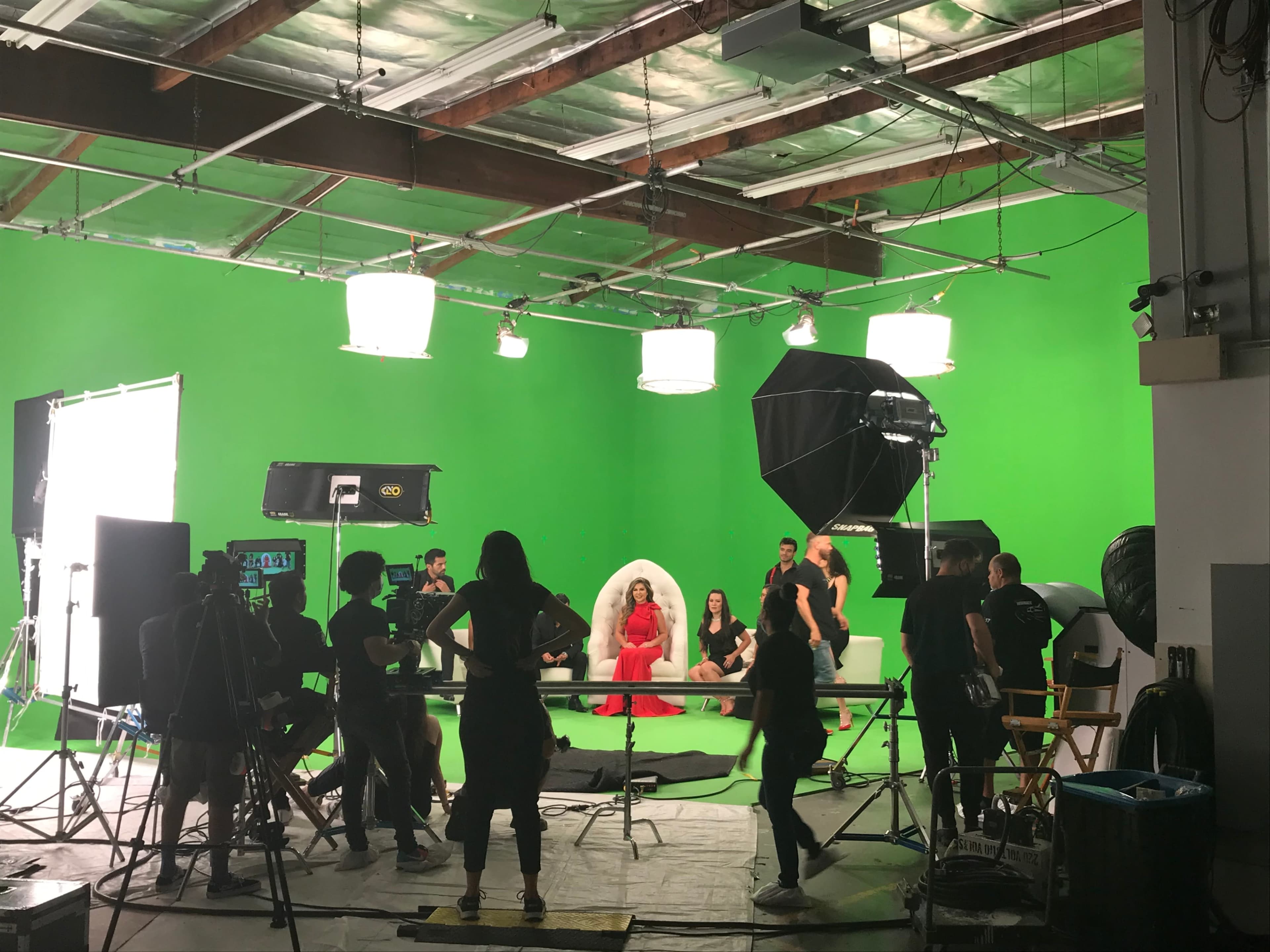 A film crew sets up in a green screen studio, featuring a central figure in a red dress surrounded by additional people and various lighting equipment.