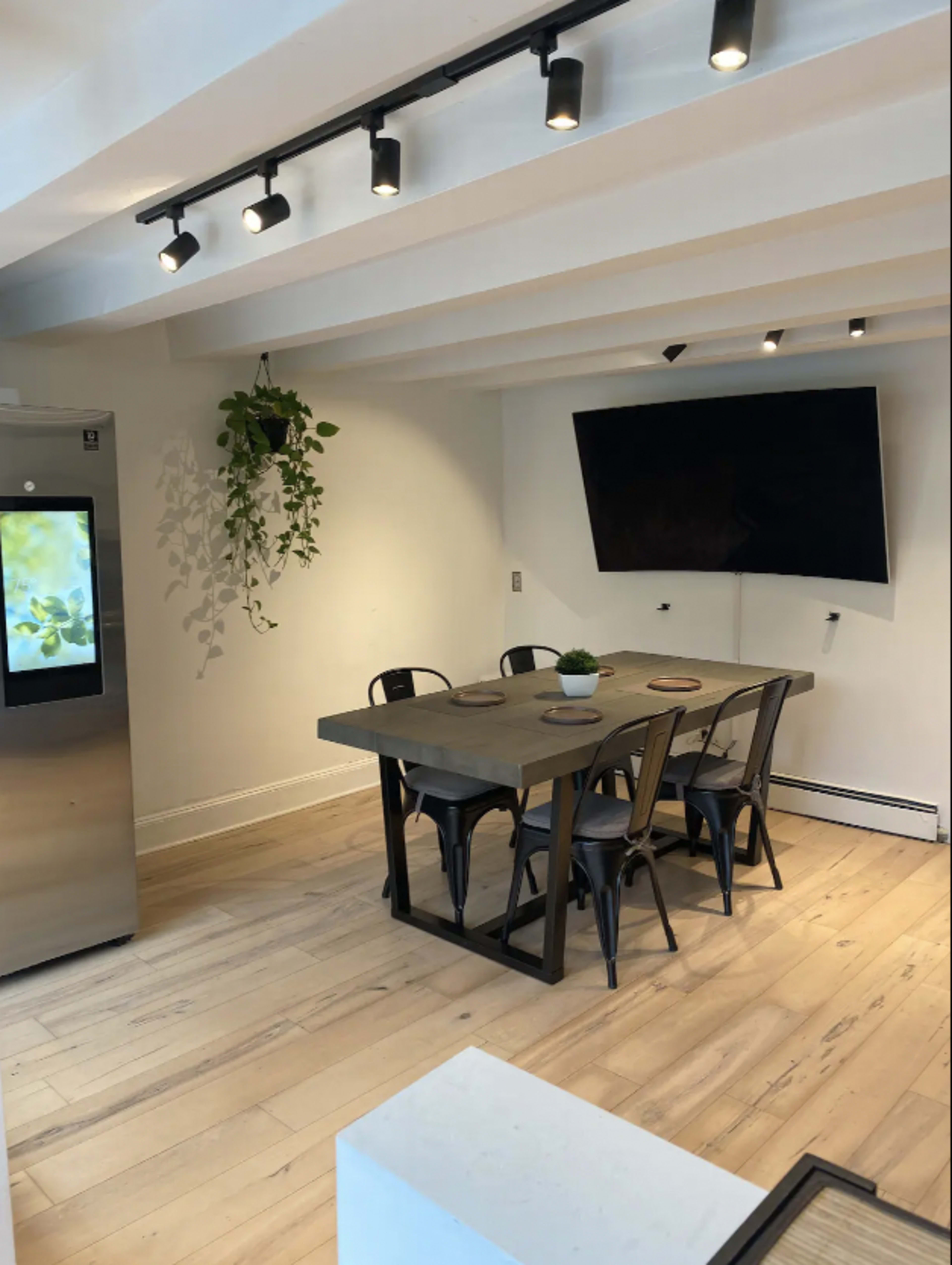 A modern dining area features a rectangular table with six chairs, a large flat-screen TV on the wall, and a refrigerator in the background.
