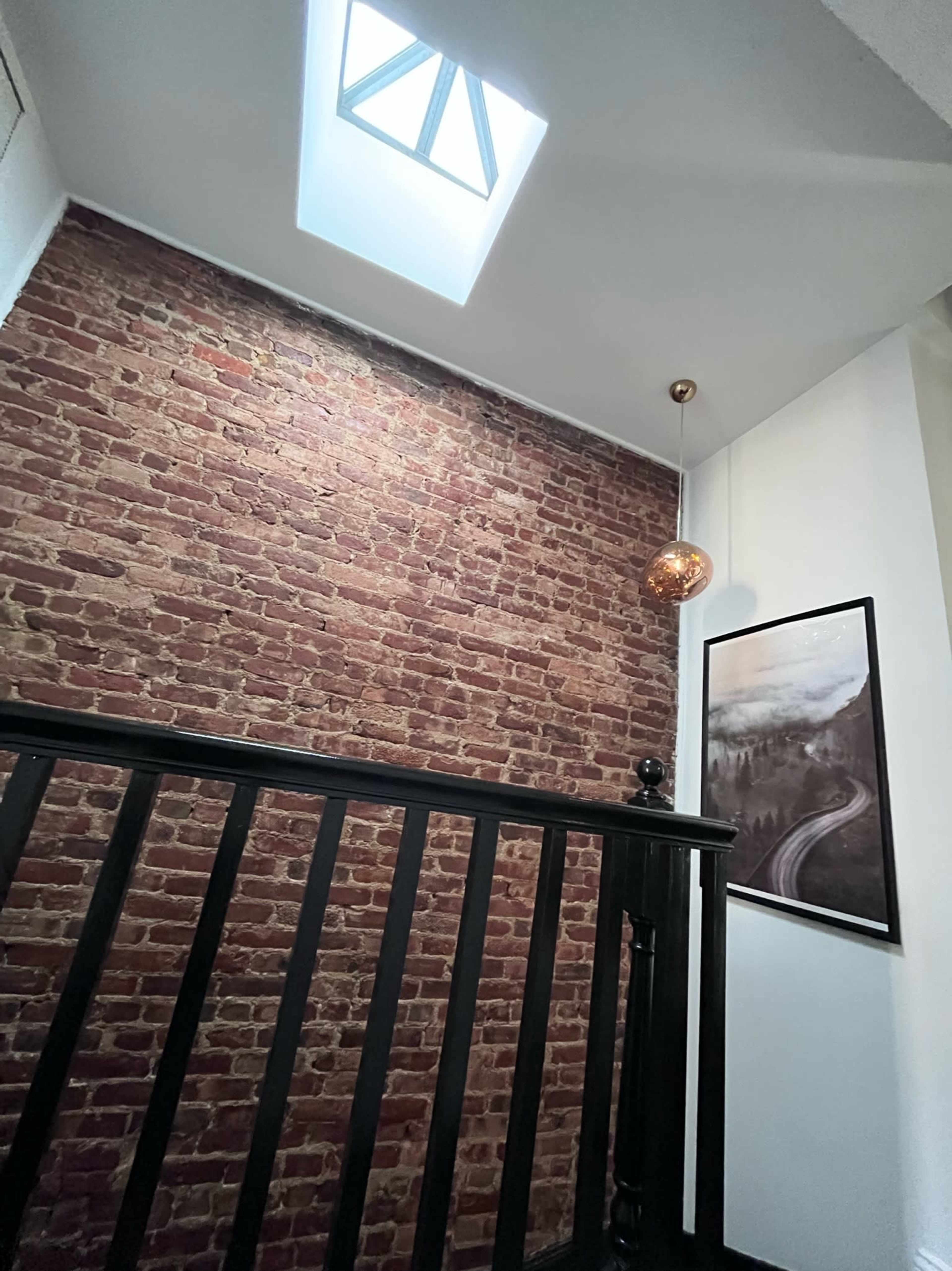The image shows a staircase area with a brick wall, a skylight overhead, and a framed picture depicting a road.