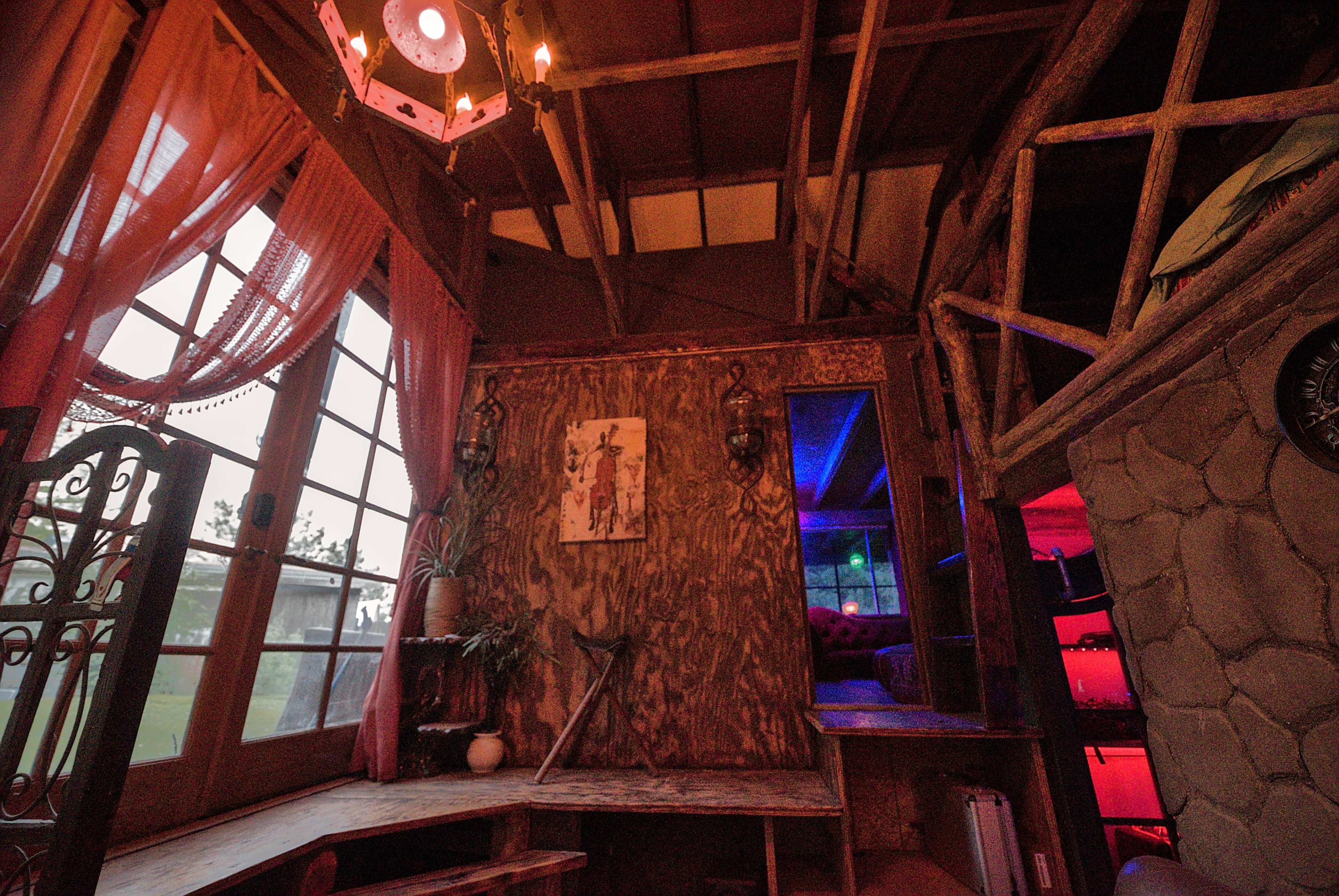The image depicts the interior of a rustic cabin featuring wooden beams, a window with red curtains, and a stone wall with ambient lighting.