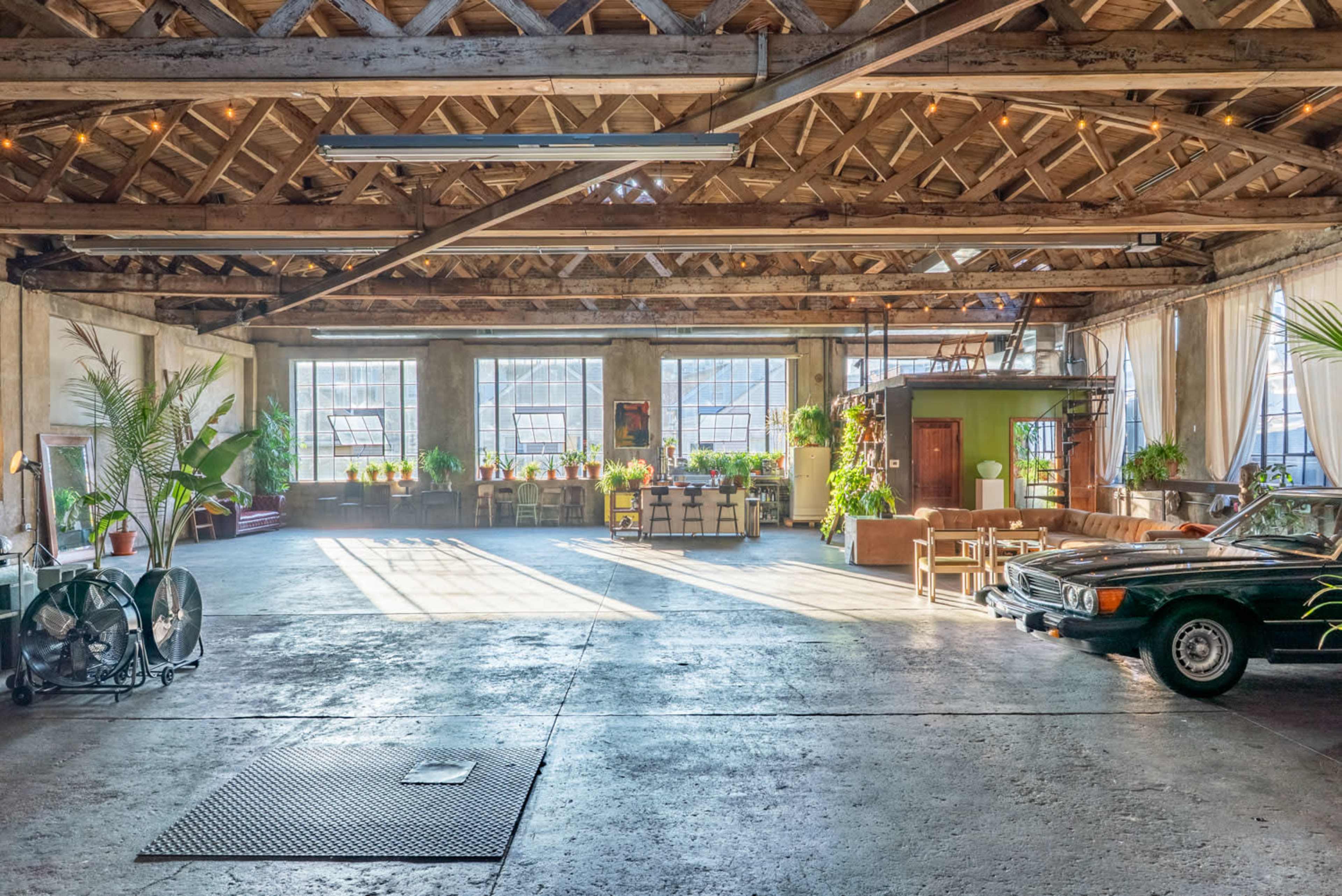 The image shows a spacious loft-style room with high wooden beams, large windows, and various indoor plants, featuring a vintage car parked on a concrete floor.