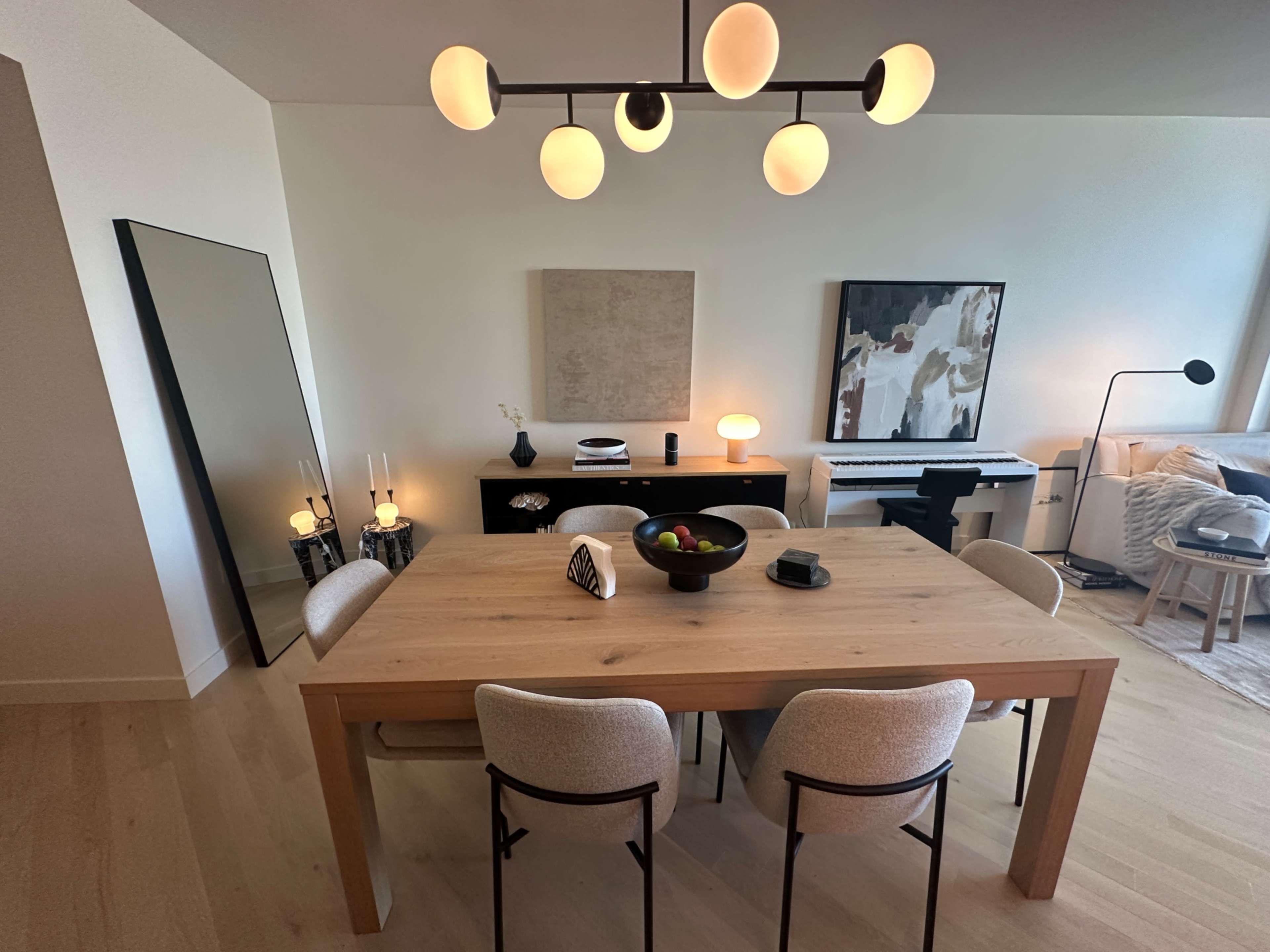A dining area features a wooden table surrounded by light-colored chairs, a large mirror, and various decorative elements on the walls and furniture.